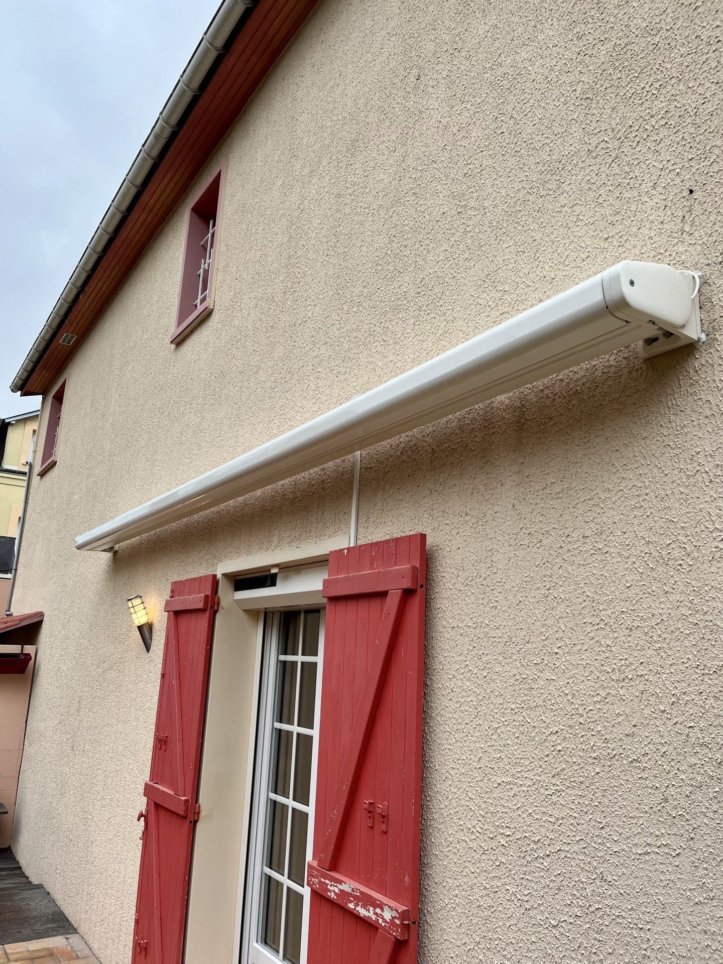Extérieur d'une maison beige avec des volets rouges et un long store blanc rétractable au-dessus d'une porte