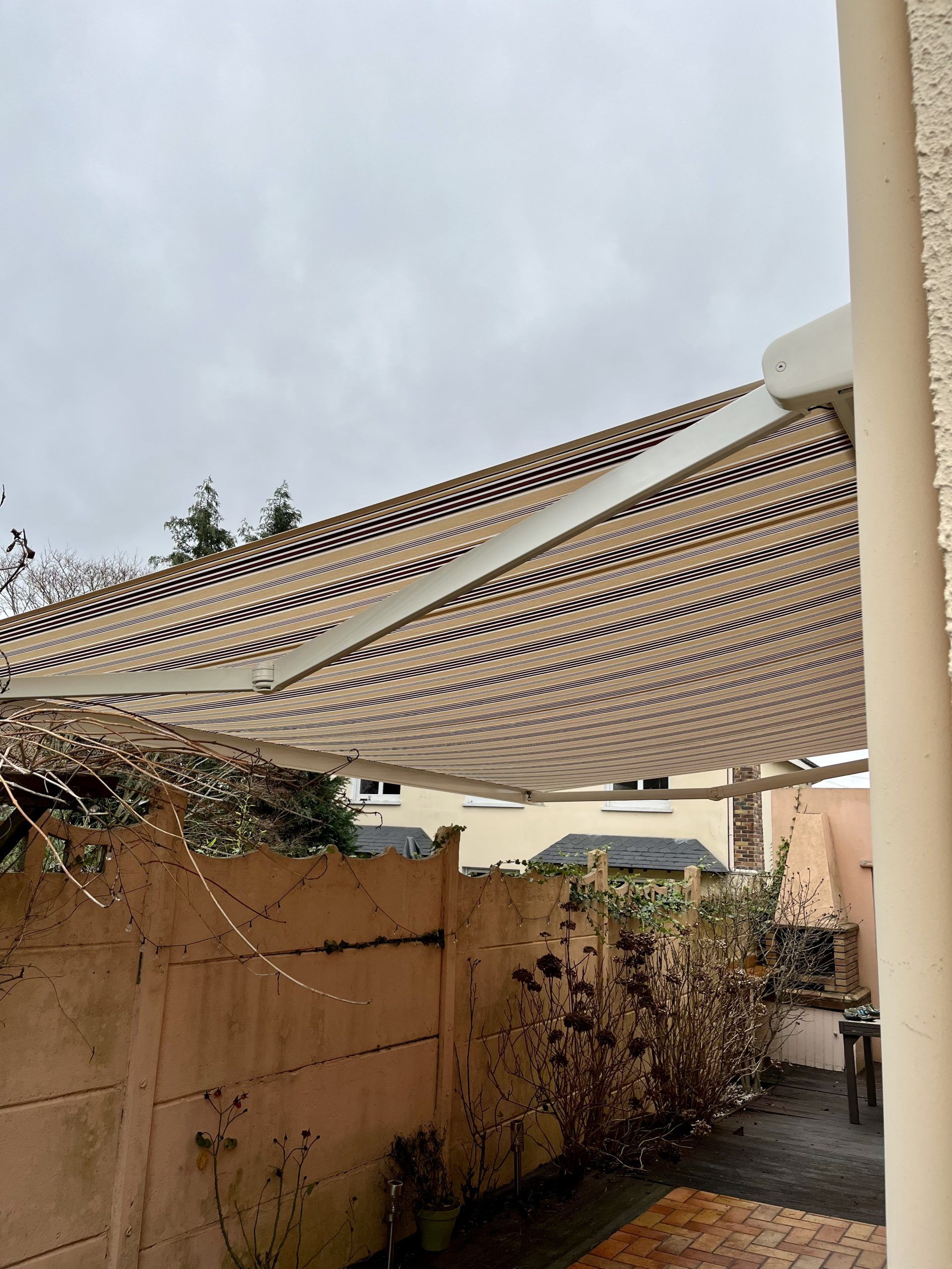 Un auvent rayé s'étendait au-dessus d'un patio étroit, bordé de murs beiges et de plantes, sous un ciel nuageux.