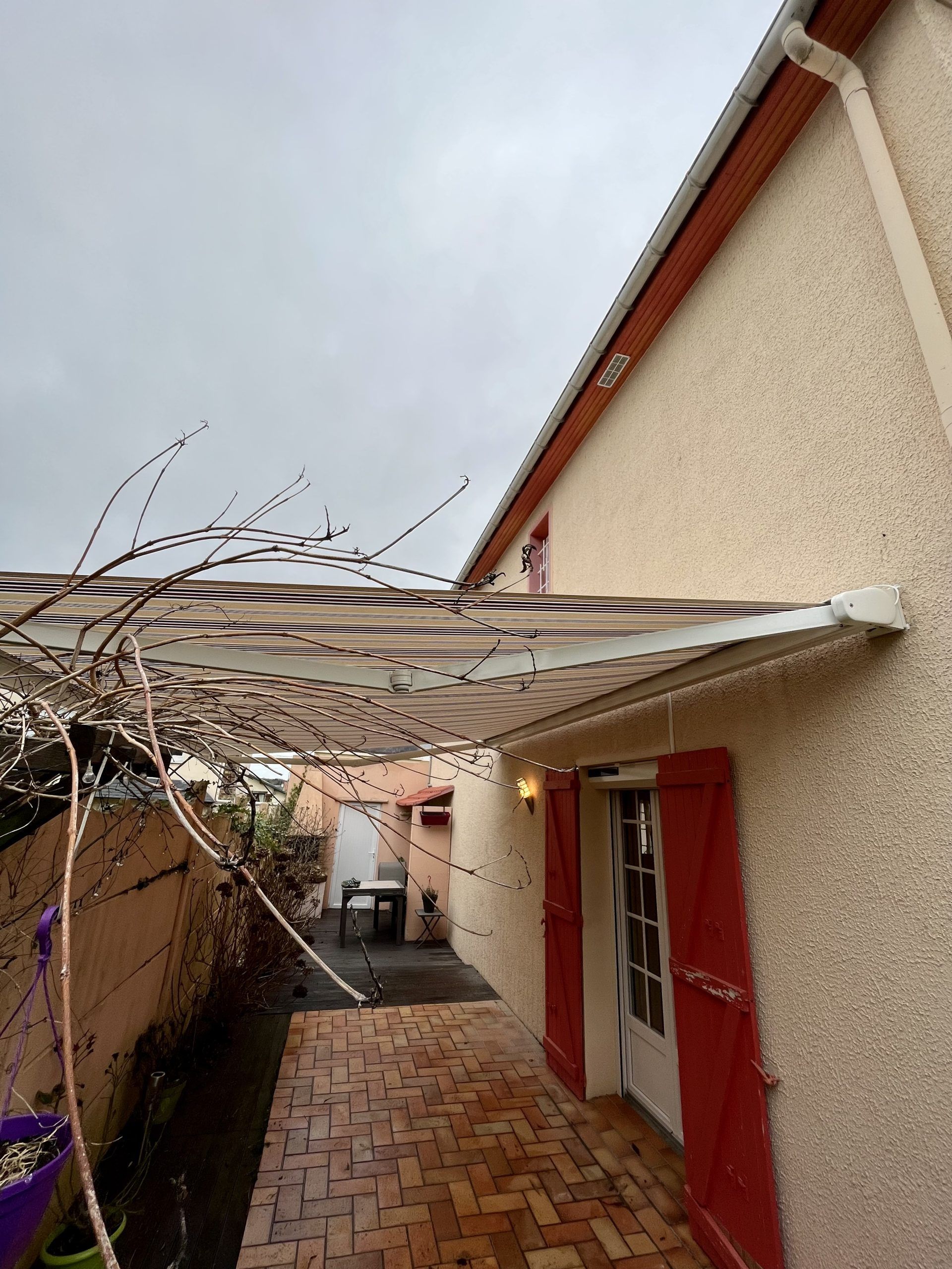 Étroite terrasse en briques jouxtant une maison beige aux portes rouges et à la pergola blanche.