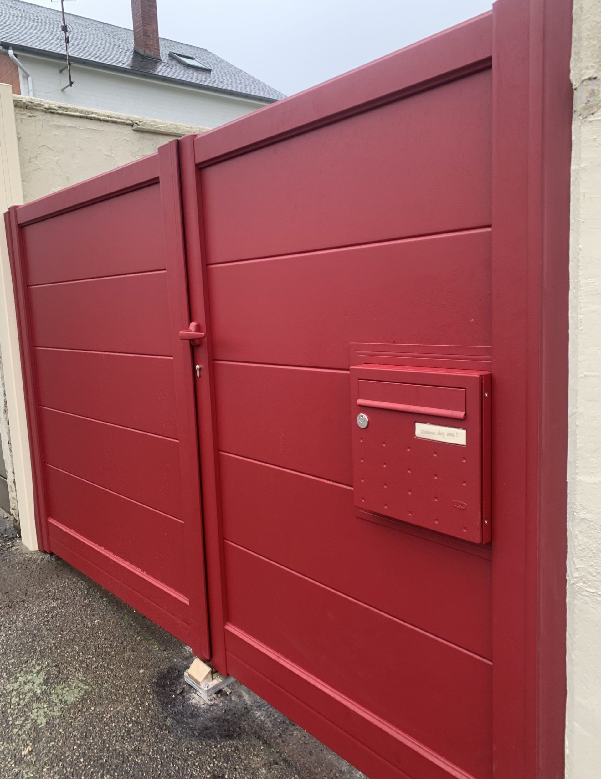 Portail métallique rouge avec porte piétonne et boîte aux lettres sur une allée de gravier.