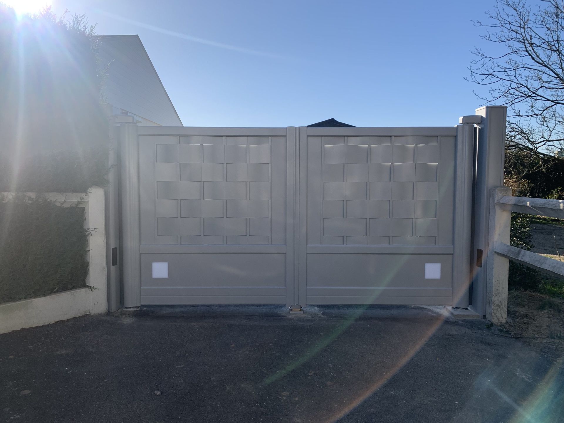 Portail double beige devant une maison, éclairé par un soleil éclatant avec un effet de halo.
