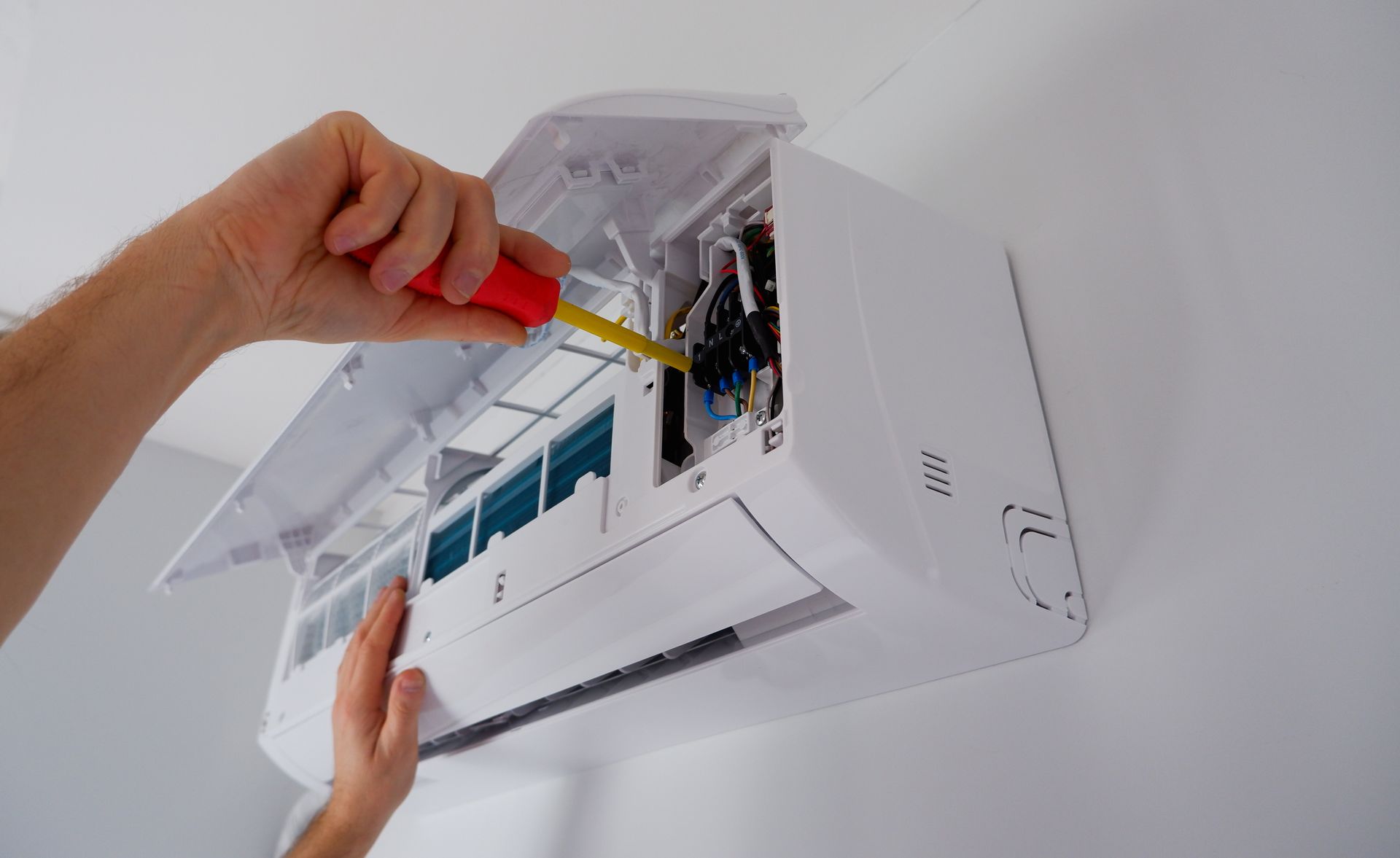 Eine Person repariert mit einem Schraubenzieher eine weisse Klimaanlage an der Wand.