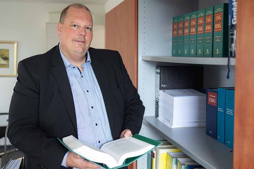 Ein Mann im Anzug hält ein Buch vor einem Bücherregal.