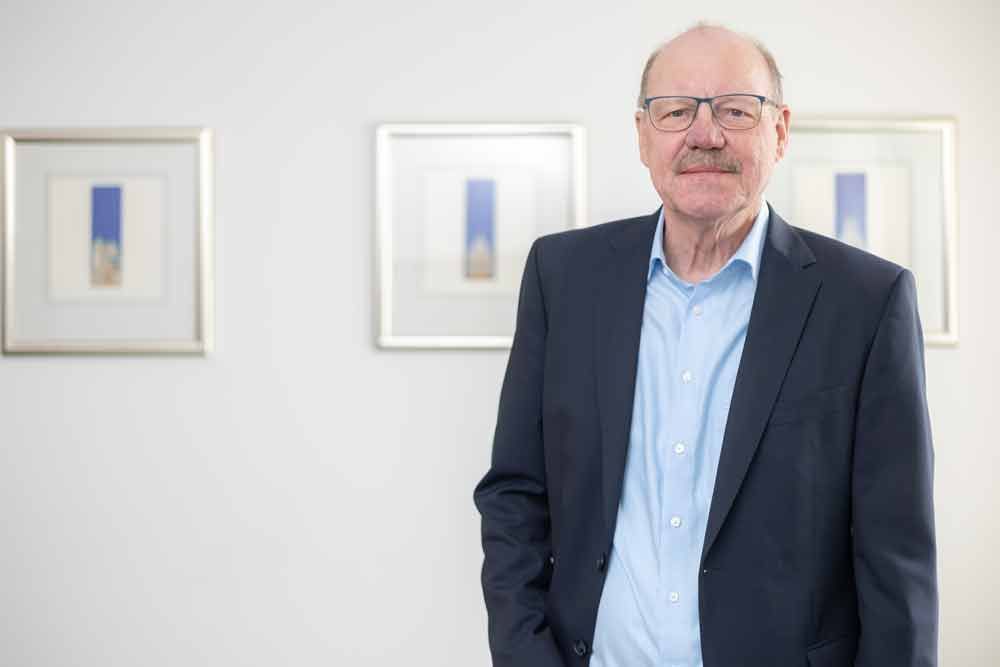 Ein Mann in Anzug und Brille steht vor einer Wand mit Gemälden.