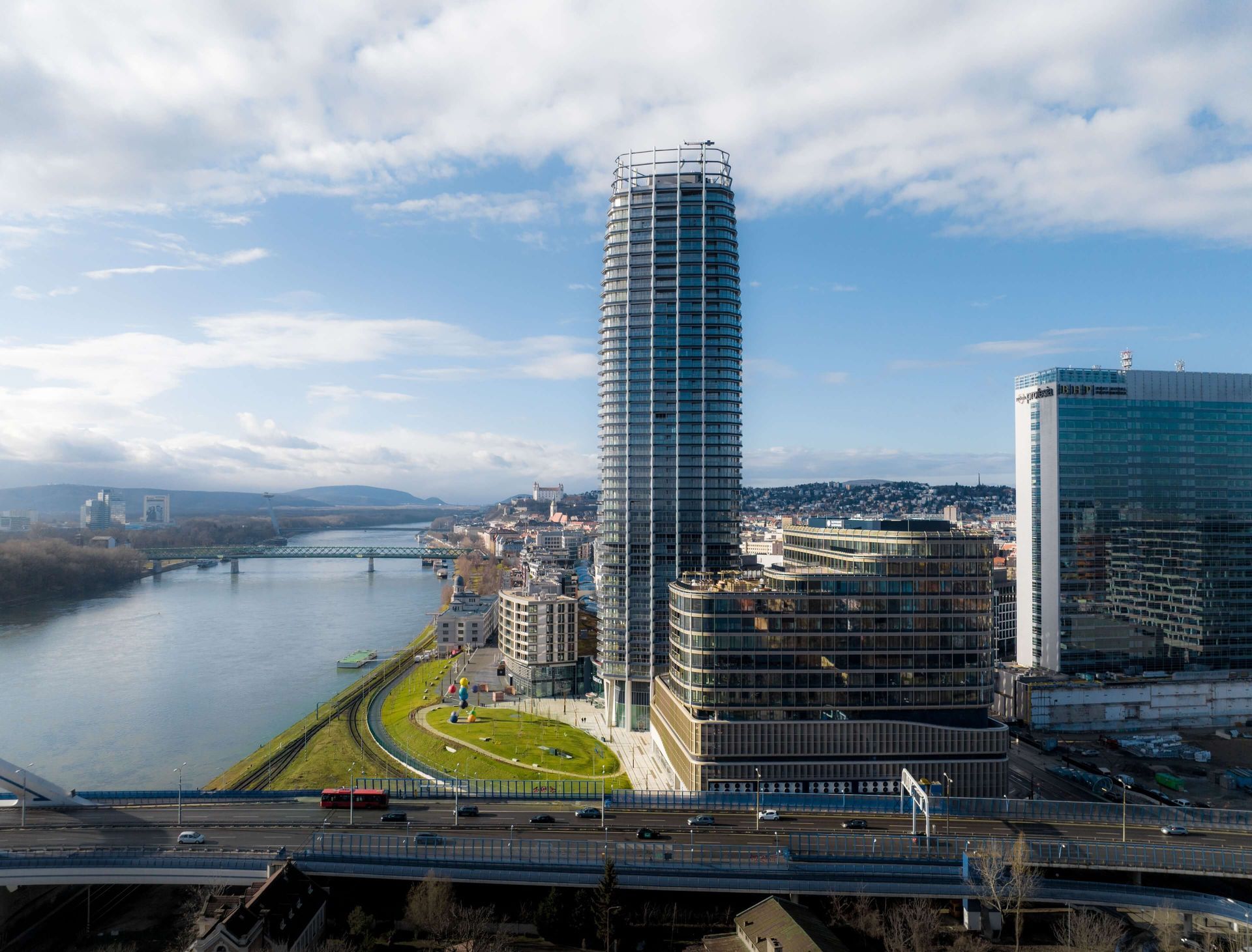 High-angle view of a skyscraper next to a river. The clear sky has scattered clouds. City buildings are in the background.