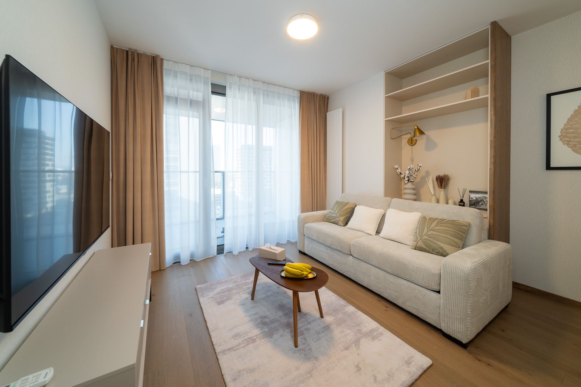 Living room with sofa, TV, built-in shelves, and window with curtains; neutral colors.