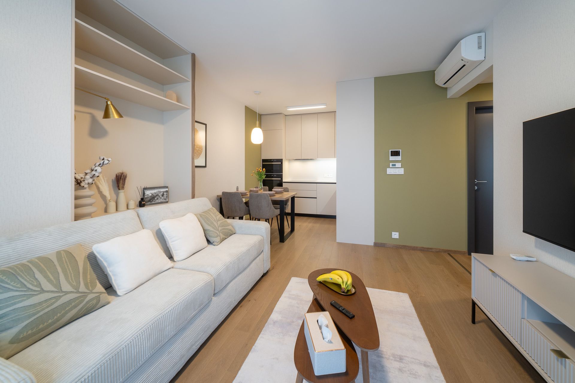 Cozy living room with a couch, coffee table, and open kitchen visible. Neutral tones with green accent wall.