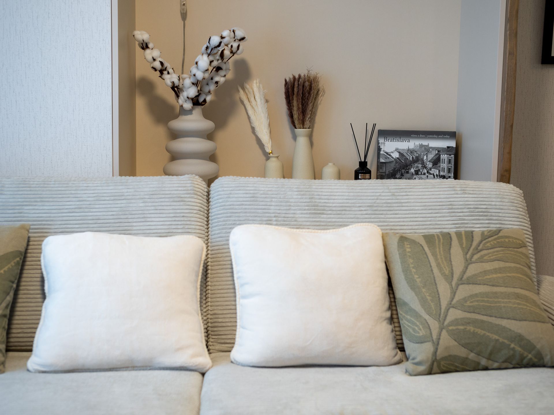 Cozy living room with beige couch, white pillows, and decorative items on a shelf: flowers, photo frame.
