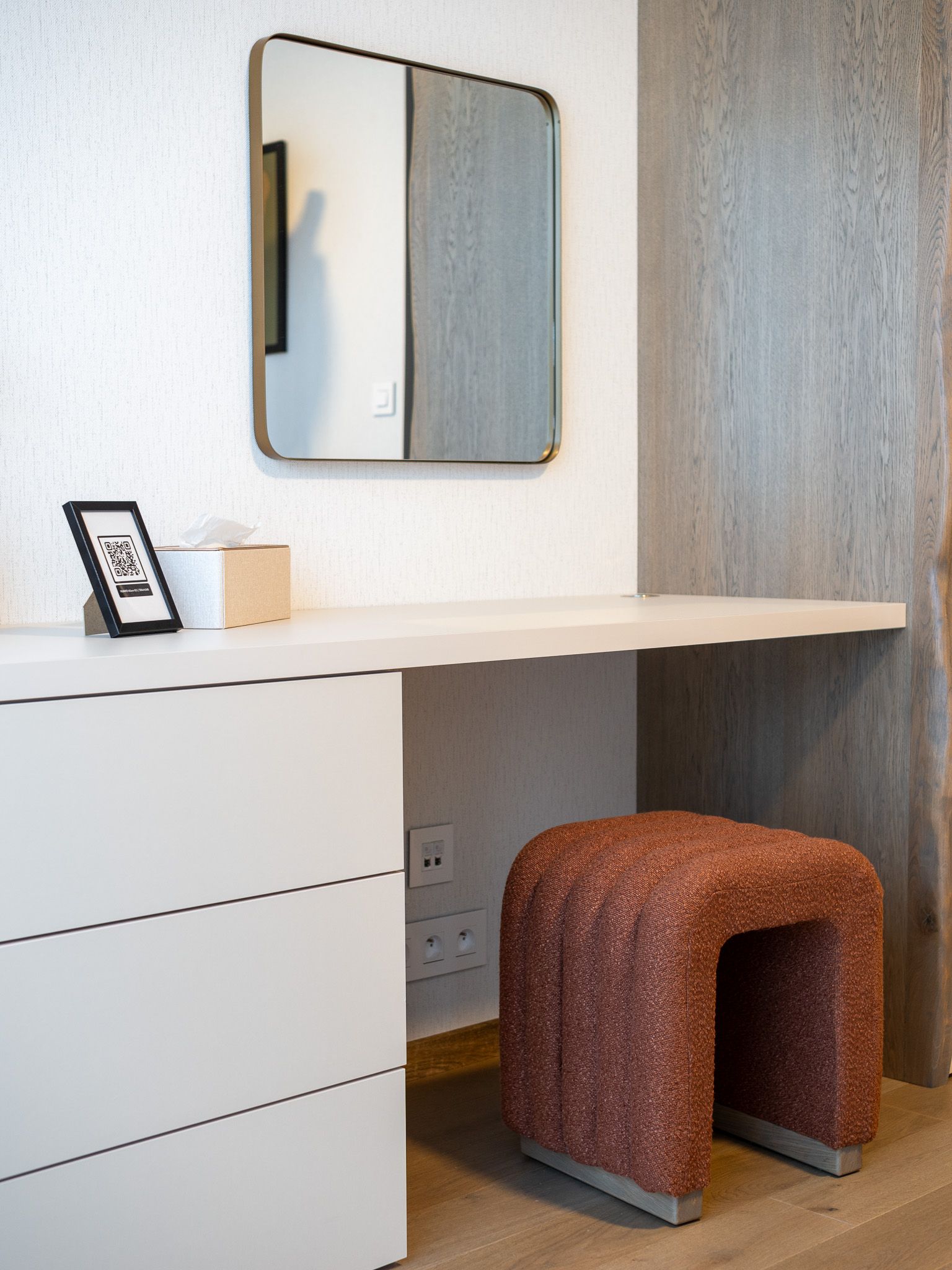 Vanity area with a mirror, built-in desk, stool, and storage drawers. Neutral colors with red stool.