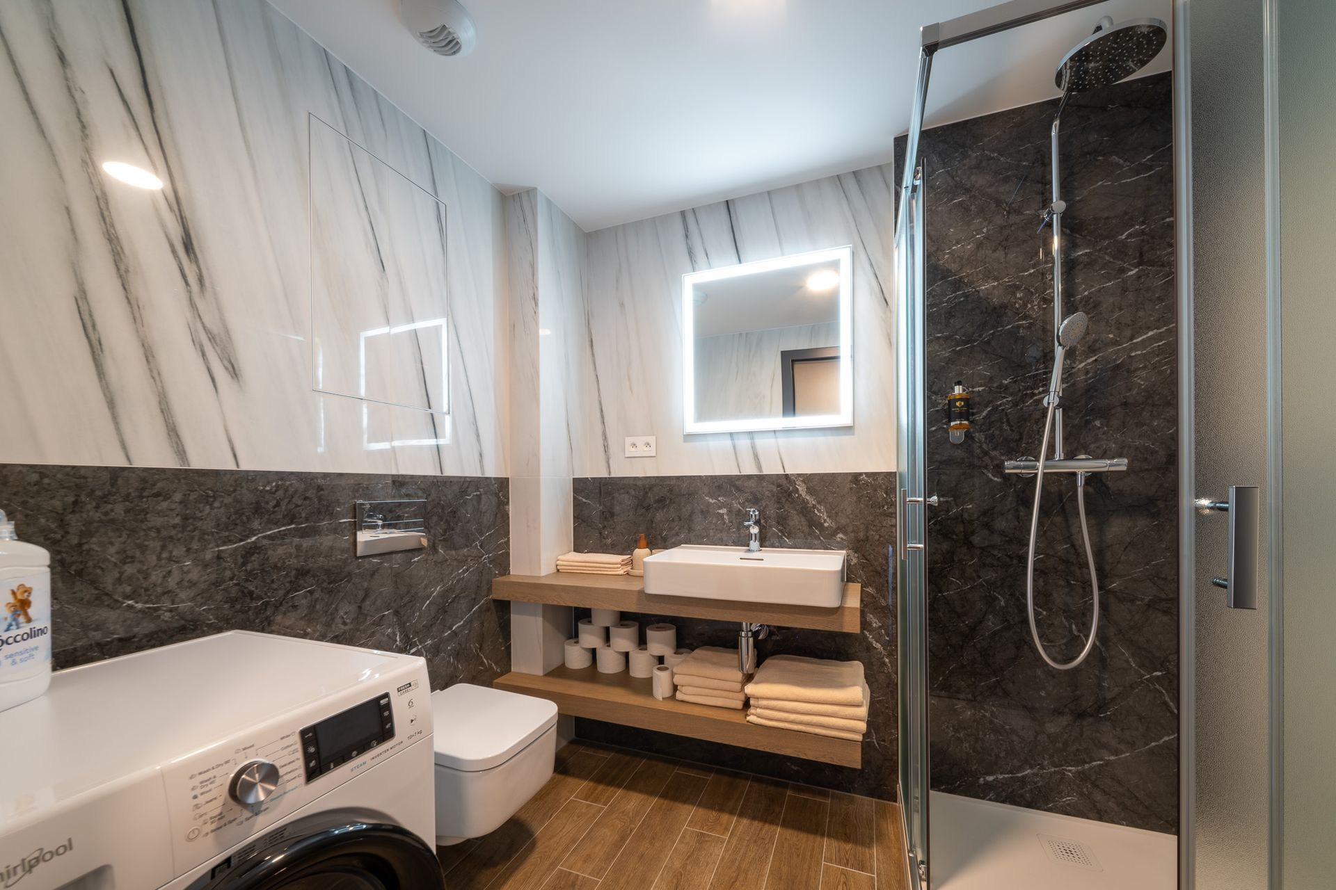 Modern bathroom with marble walls, shower, toilet, sink, washing machine, and wooden shelves with towels.
