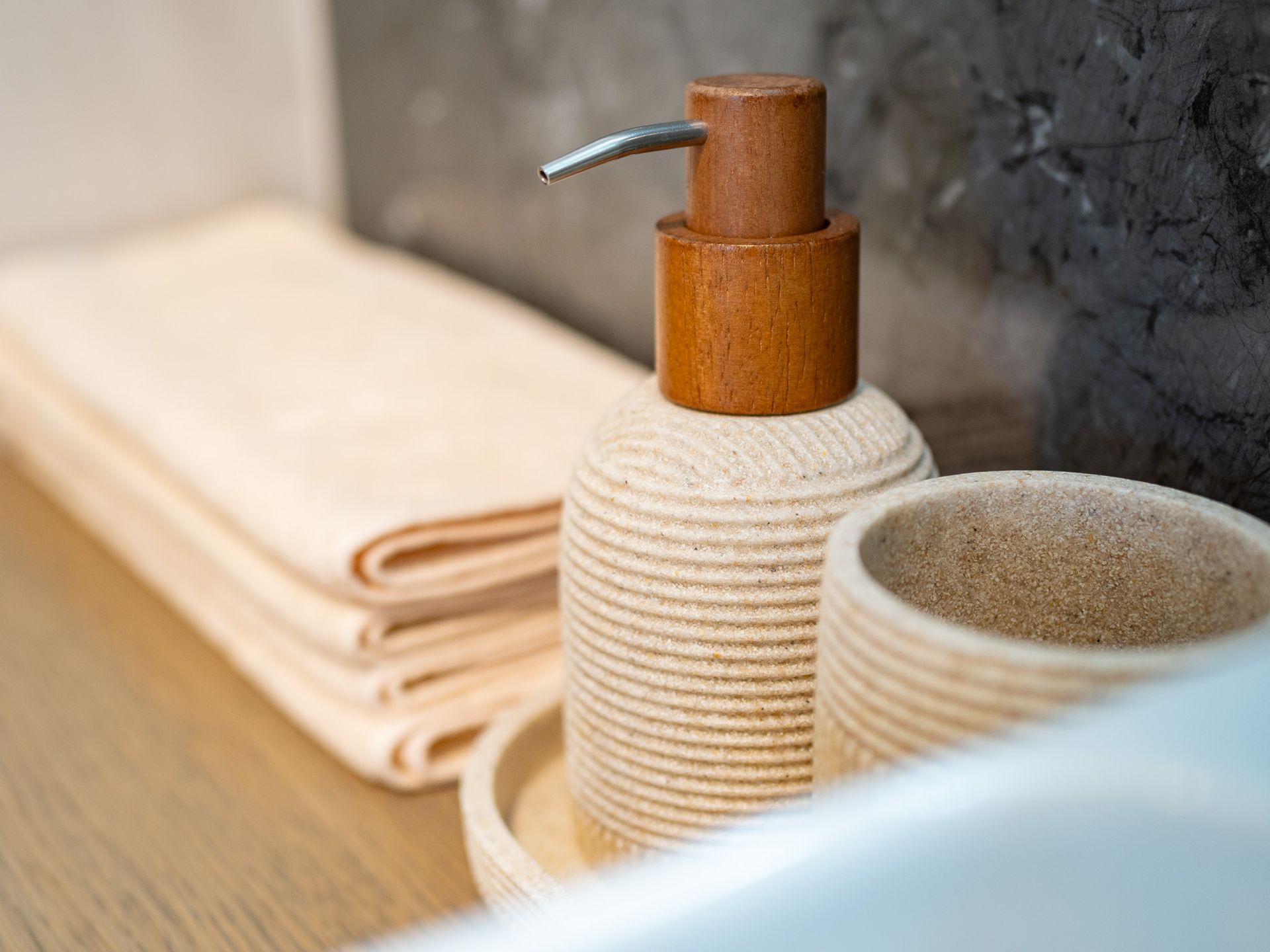 Soap dispenser, cup, and folded towels on a wooden tray, near a dark stone wall.