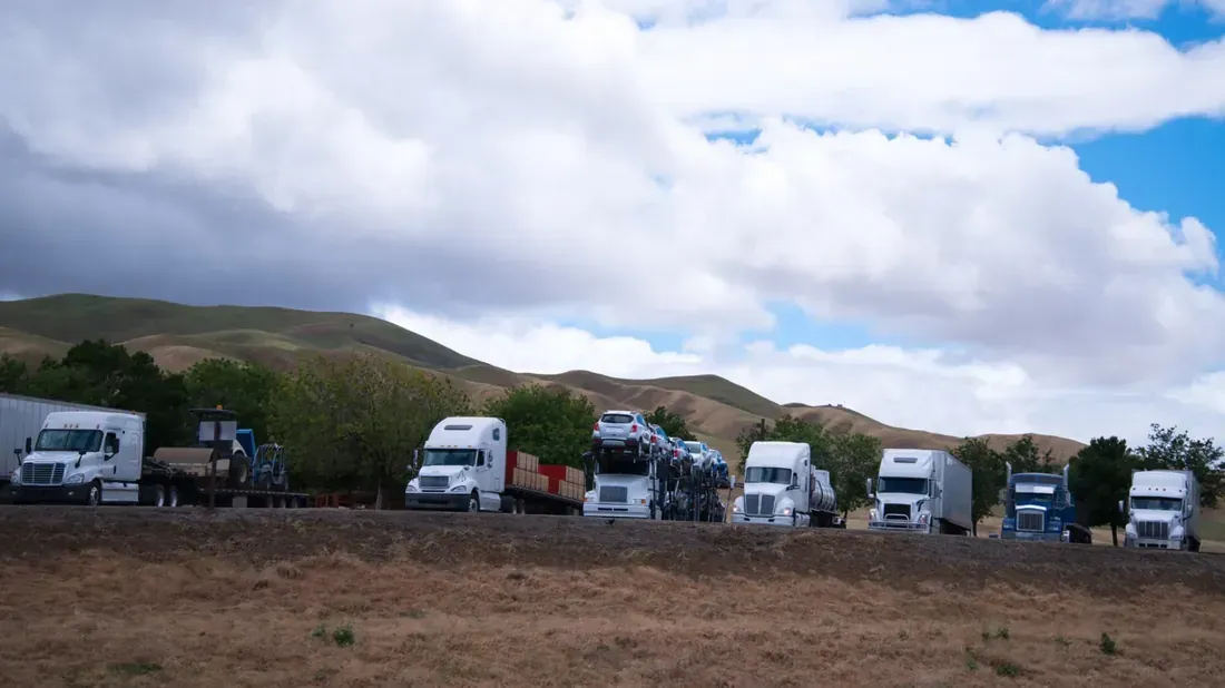 Fila de semirremolques estacionados al costado de la carretera junto a colinas marrones y cielo nublado.