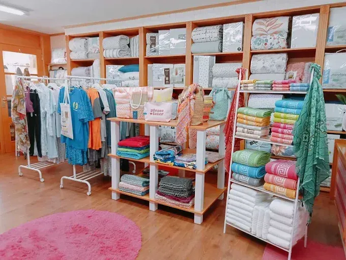 Boutique de ropa luminosa con coloridas sábanas dobladas, vestidos colgados y una alfombra rosa sobre un suelo de madera.