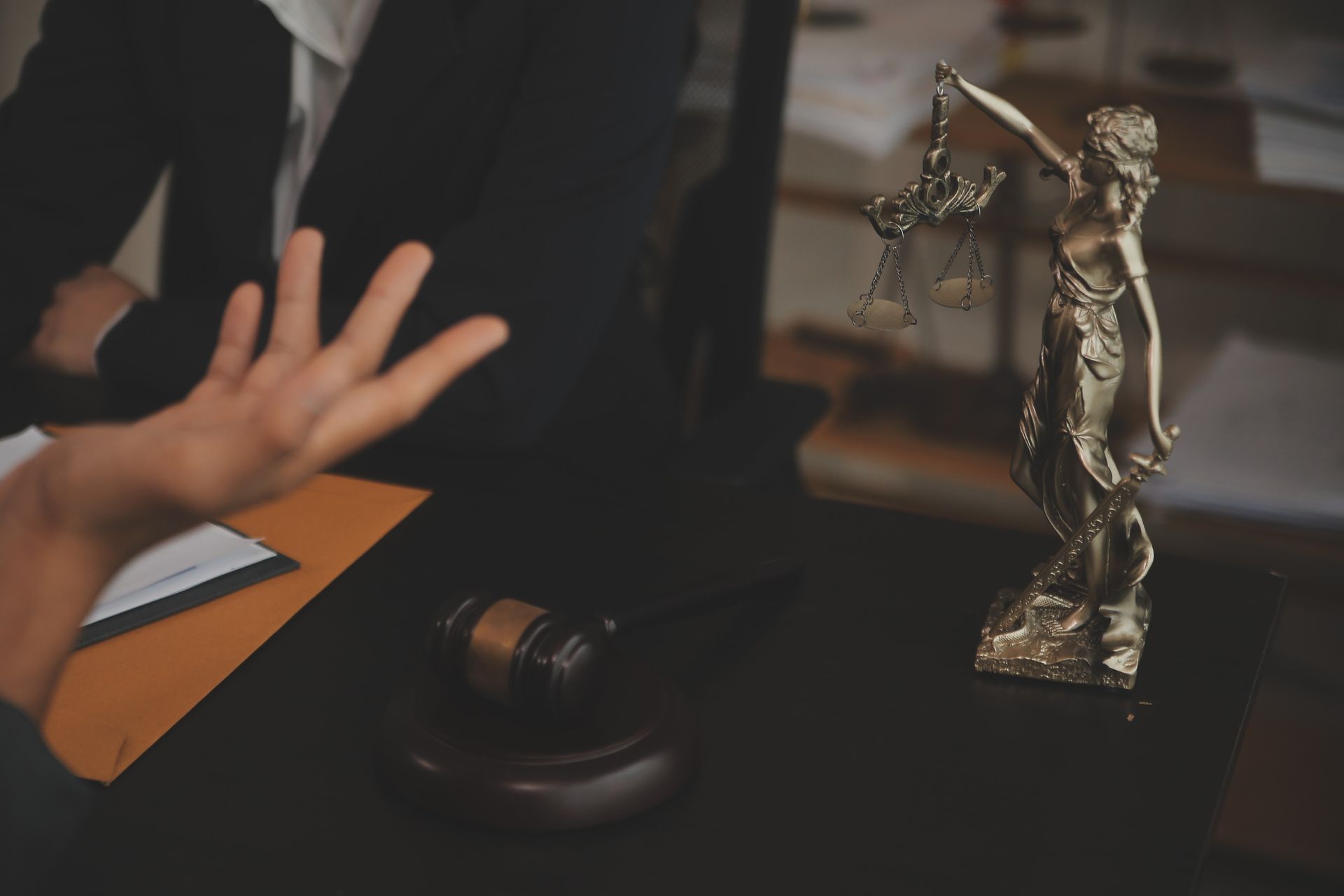 Una persona hace gestos frente a una estatua de una balanza de la justicia, un mazo y documentos sobre un escritorio, posiblemente en una sala de audiencias.