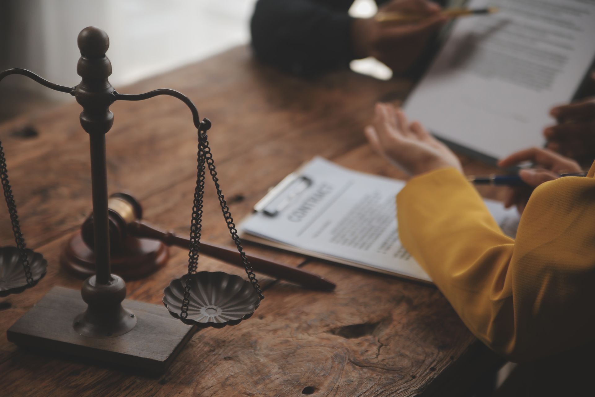 Balanza de la justicia, mazo y documentos sobre una mesa de madera con gente discutiendo papeleo.