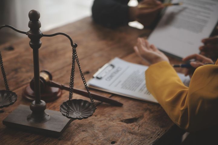 Balanza de la justicia, mazo y documentos sobre una mesa de madera con gente discutiendo papeleo.