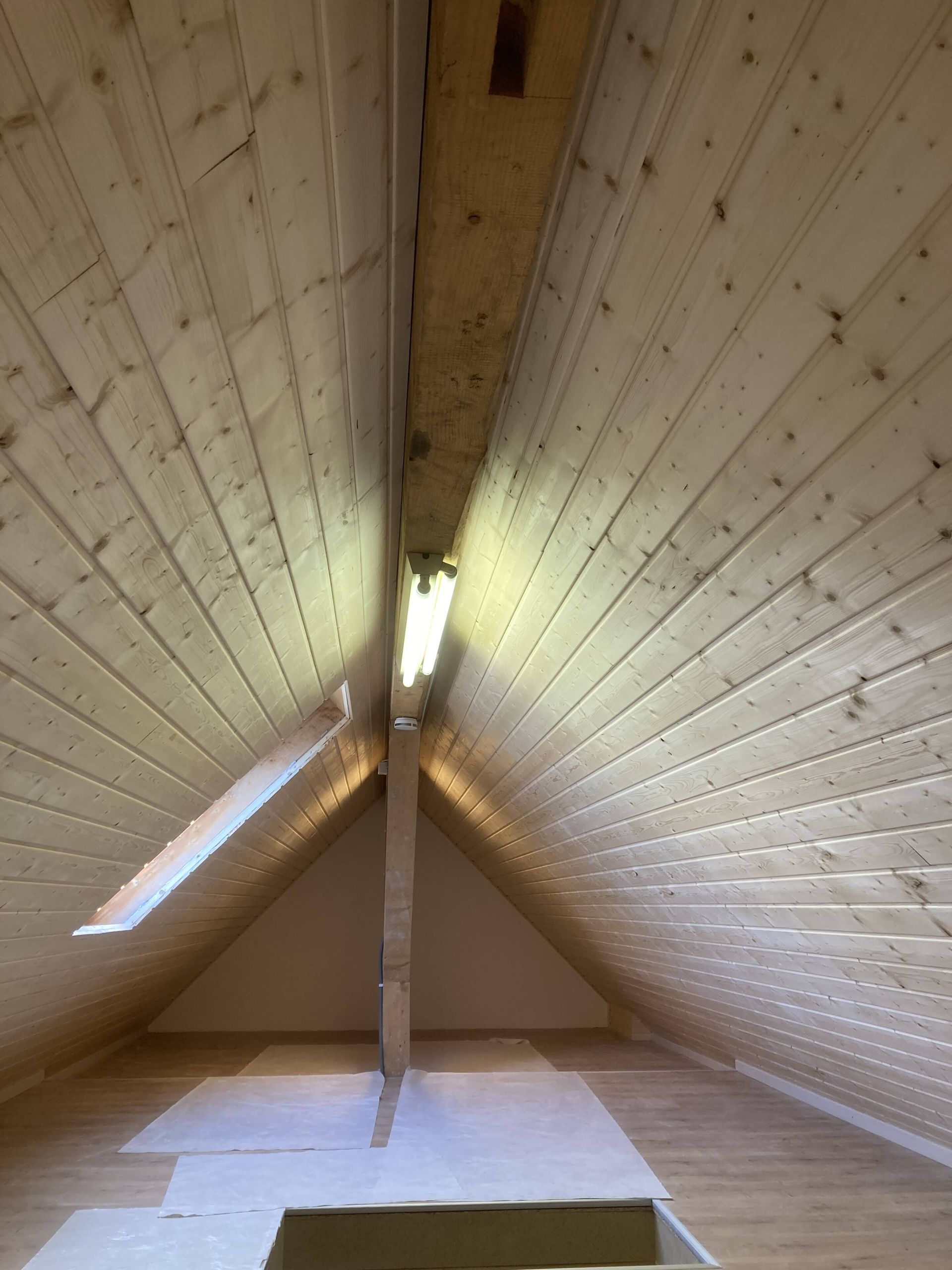 Dachzimmer mit heller Holzvertäfelung an Wänden und Decke, kleinem quadratischen Fenster und zentralem Stützbalken.