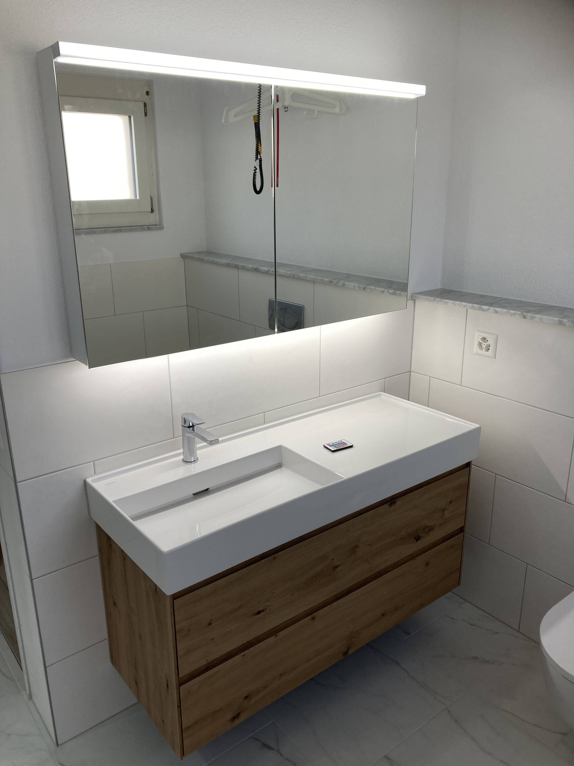 Badezimmer mit weißem Waschbecken und Spiegel, Holzschrank und heller Beleuchtung.