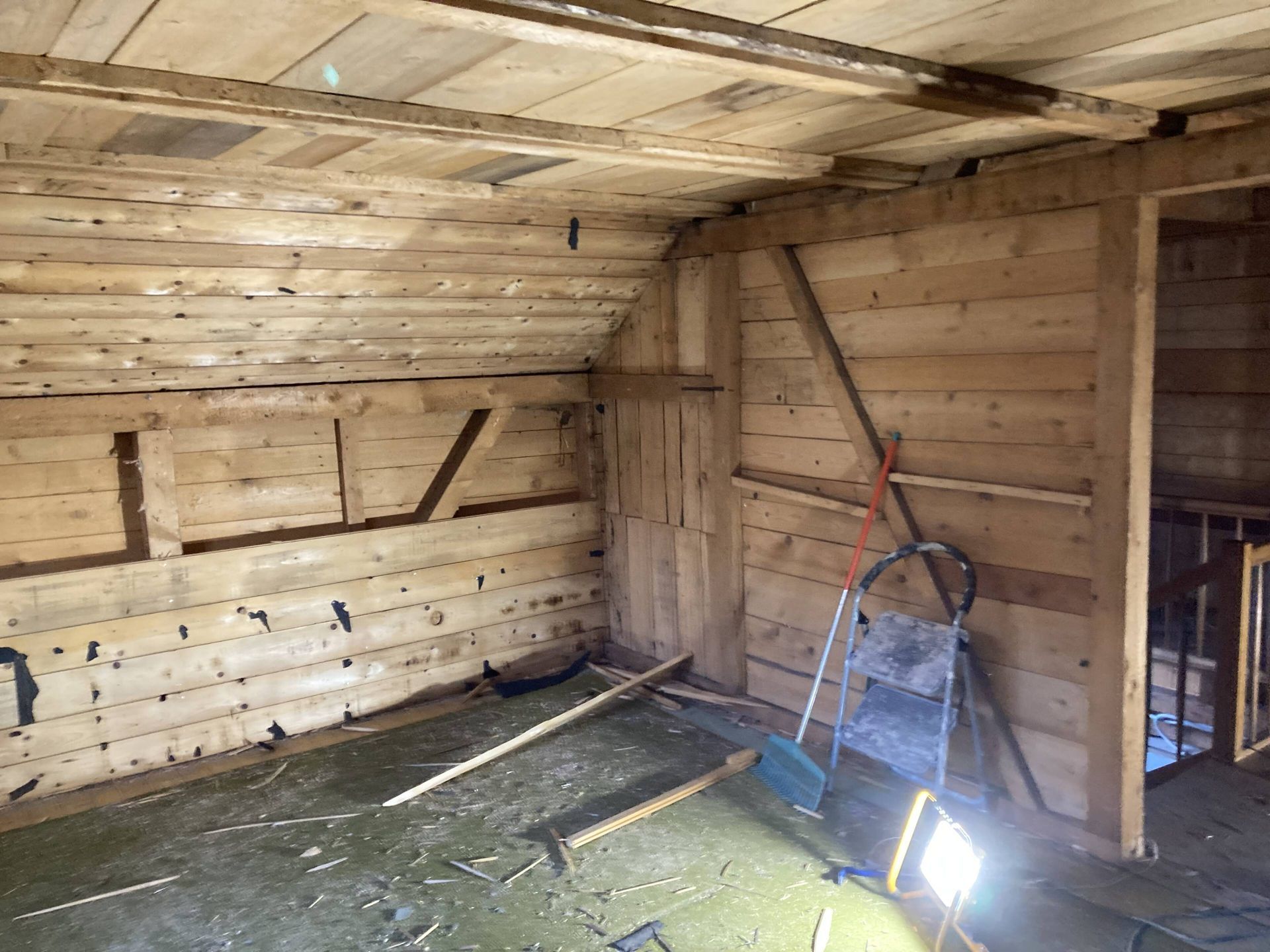 Dachbodenraum mit Holzwänden; eine Leiter lehnt an der Wand. Verstreute Werkzeuge und Schutt auf dem Boden.
