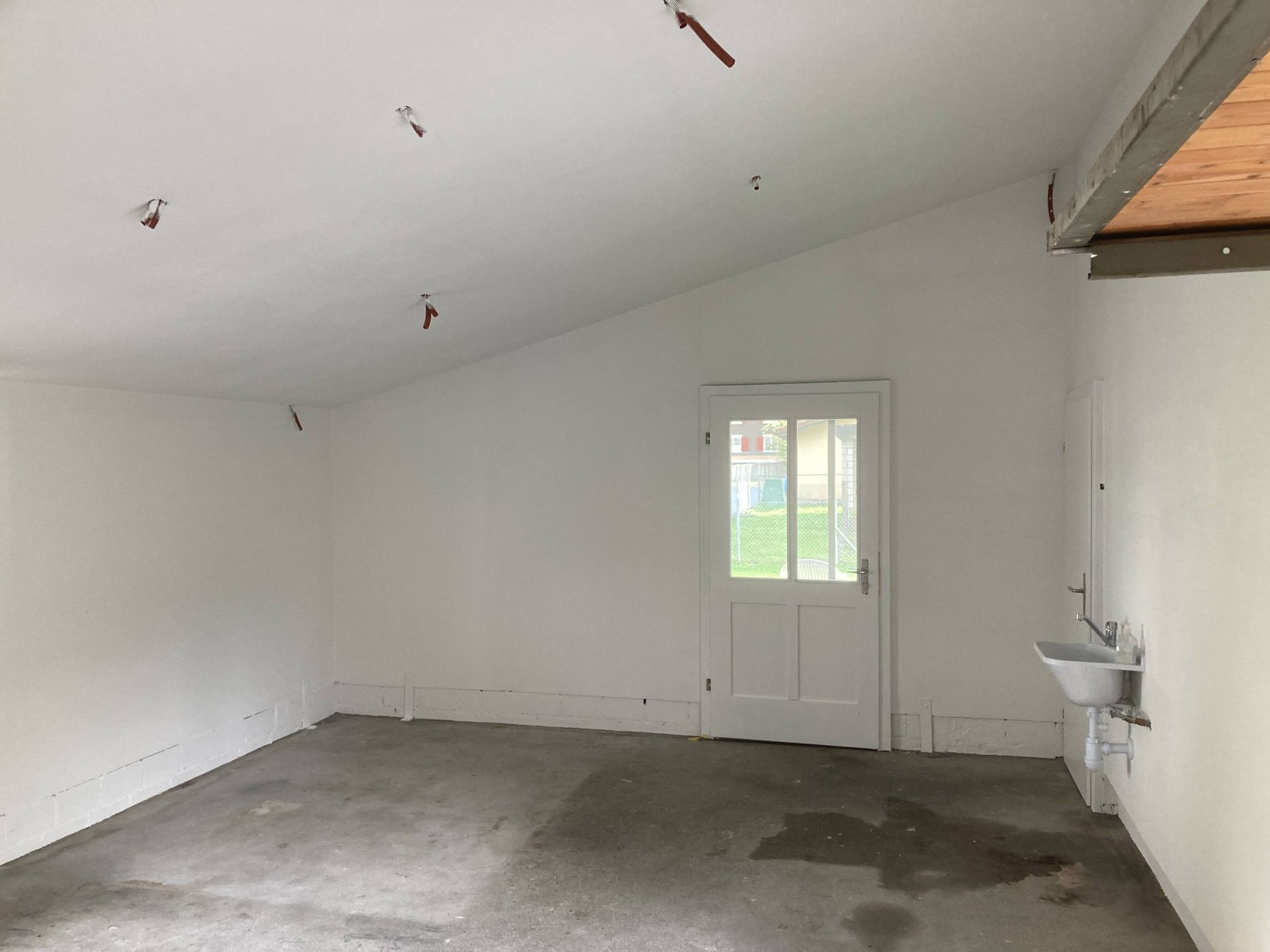 Raum mit weißen Wänden, Betonboden, Tür und Waschbecken. Über der Decke verlaufen mehrere freiliegende Kabel.