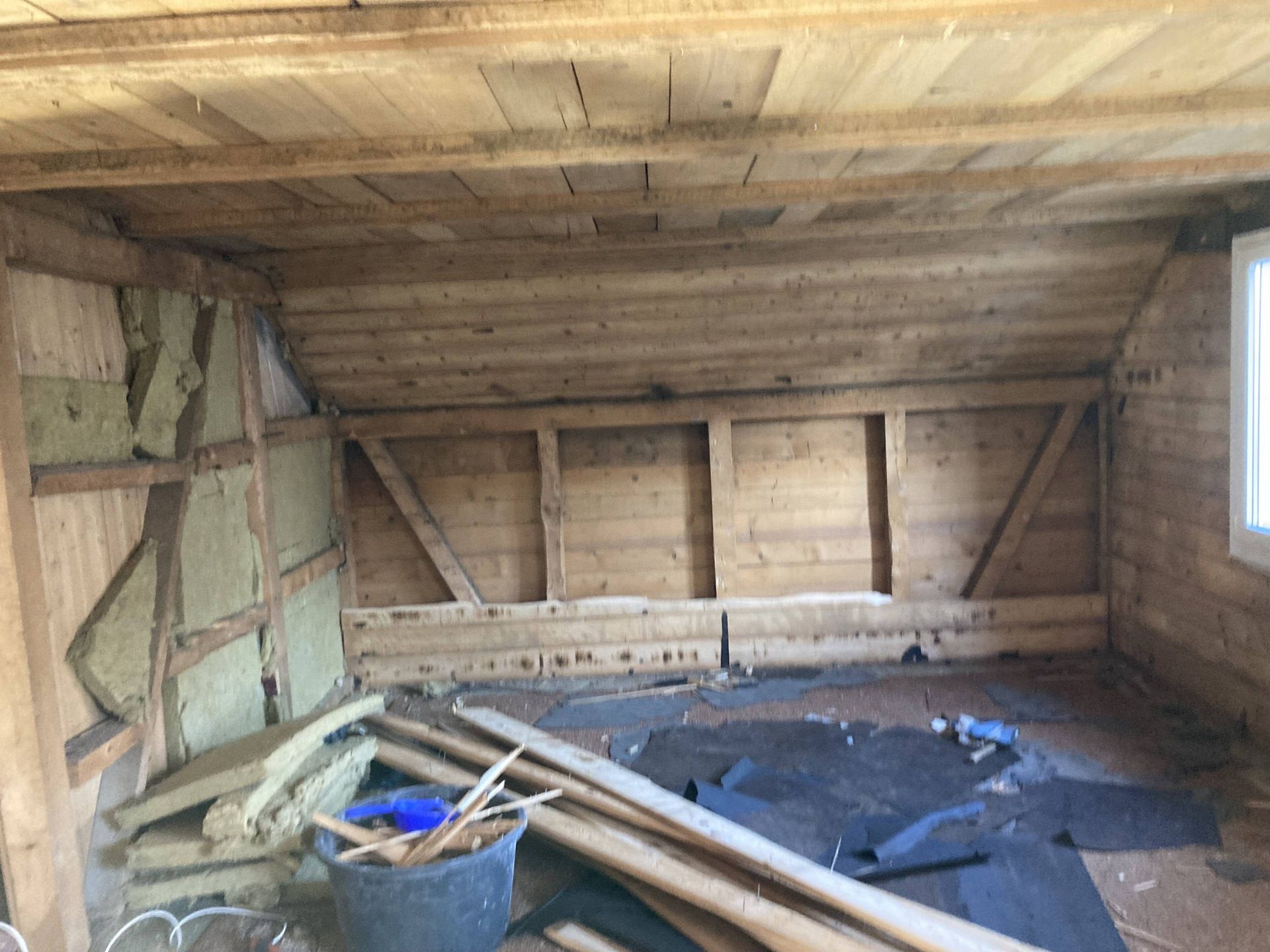 Dachzimmer mit freiliegenden Holzbalken, teilweiser Isolierung und Schutt auf dem Boden.