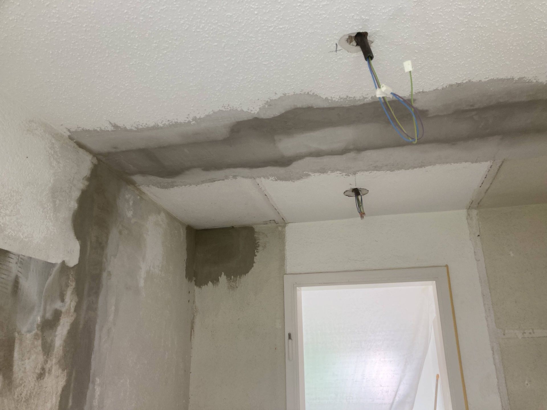 Decke und Wand im Bau mit freiliegenden Leitungen und Türöffnung. Zement und weiße Farbe sichtbar.