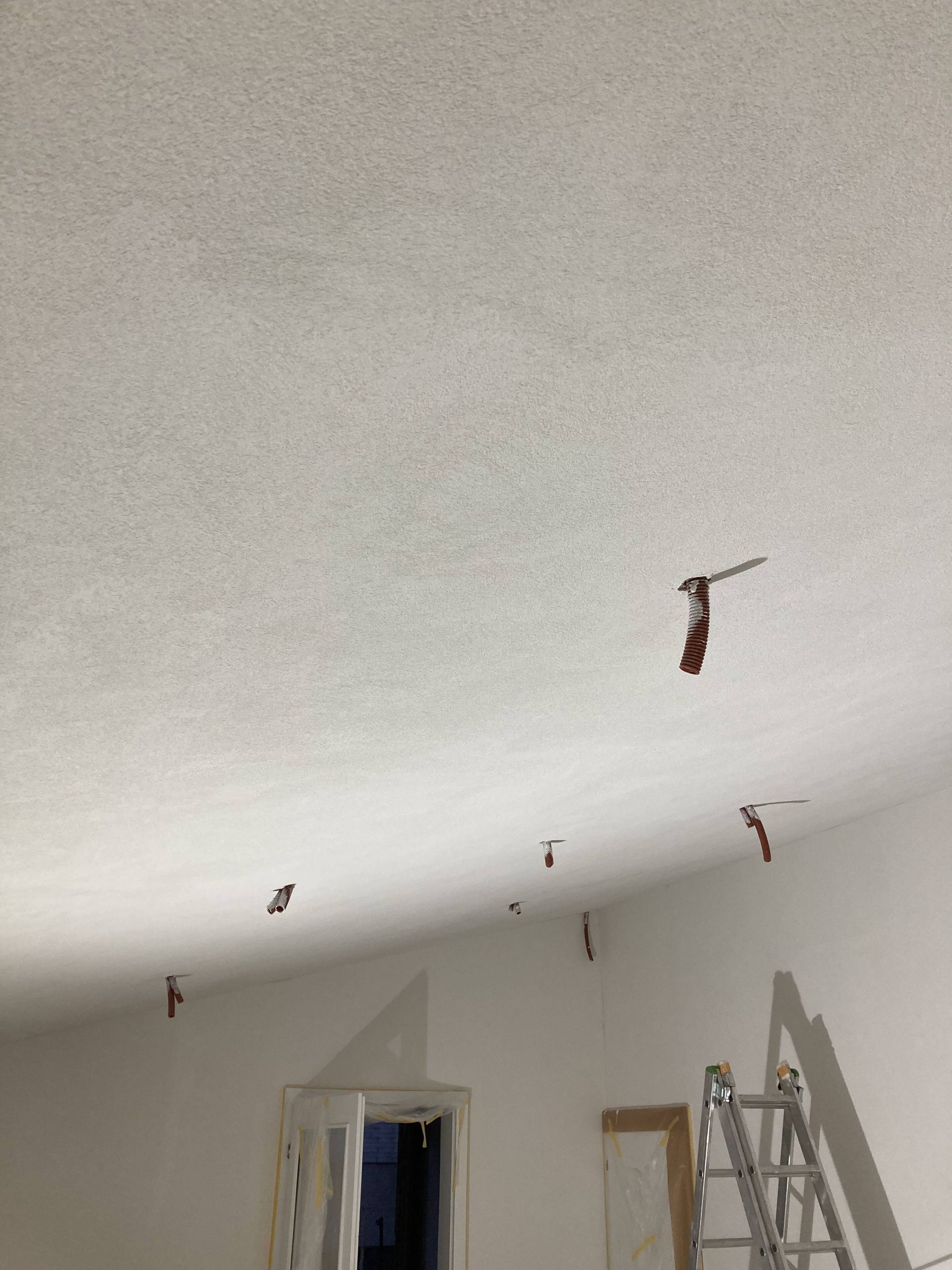 Weiße Decke mit freiliegenden Kabeln, beleuchtet durch natürliches Licht. Eine Leiter lehnt an einer weißen Wand.