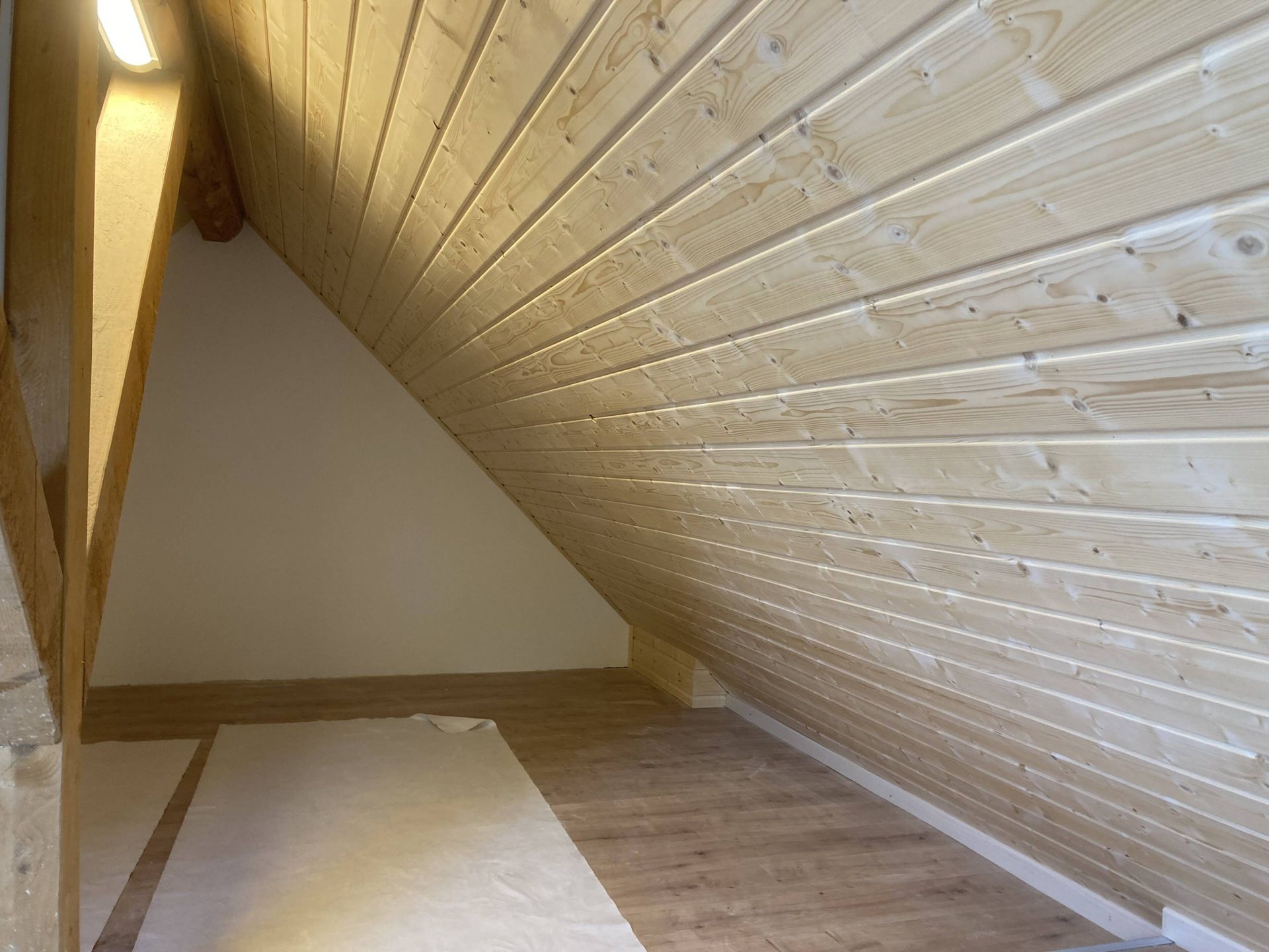 Dachzimmer mit holzgetäfelter Decke und Wänden, die zum Boden hin geneigt sind. Helles Holz, weiße Farbe und Hartholzboden.
