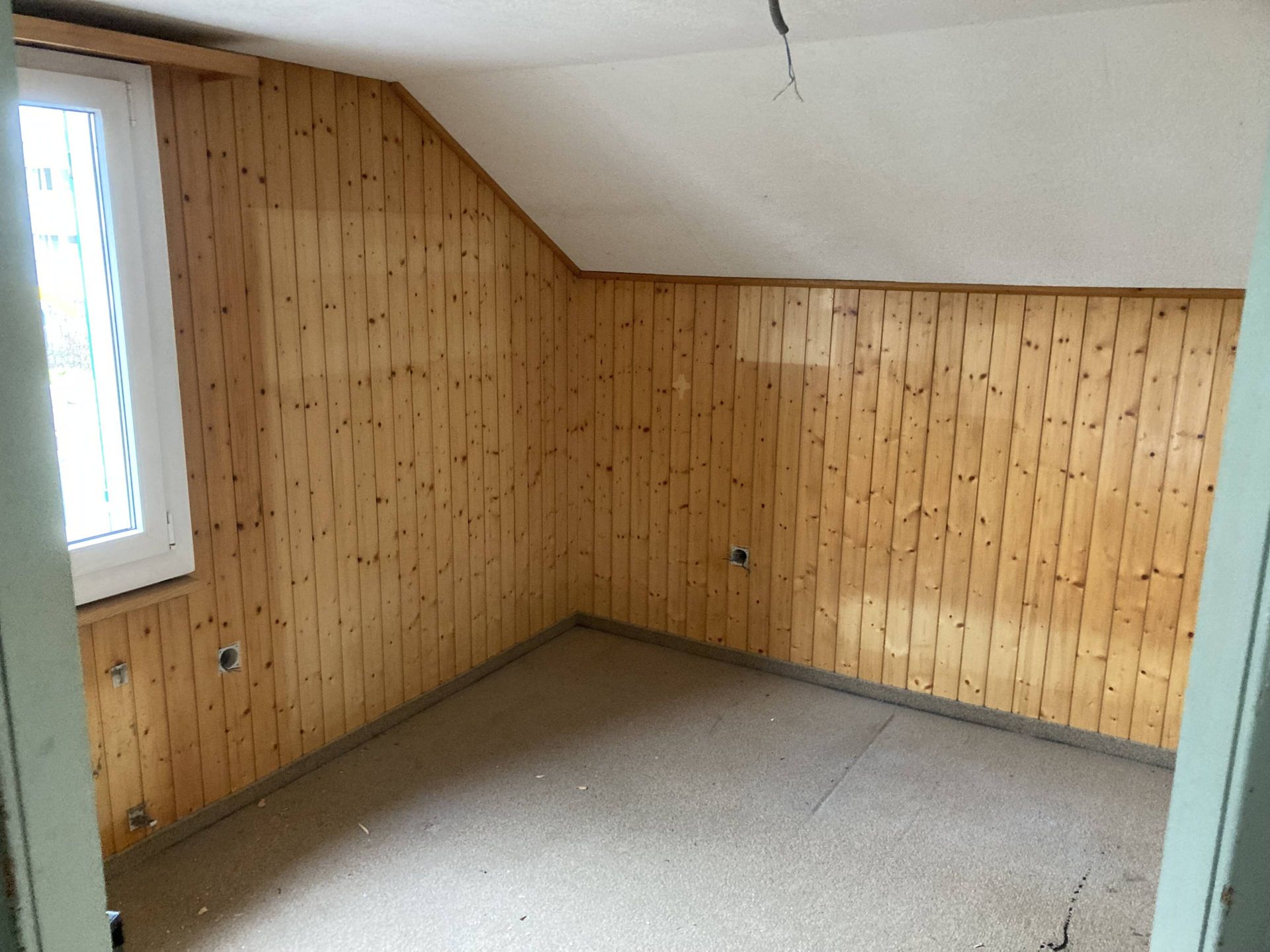 Leerer Raum mit Holzvertäfelung an den Wänden und schräger Decke. Fenster auf der linken Seite.