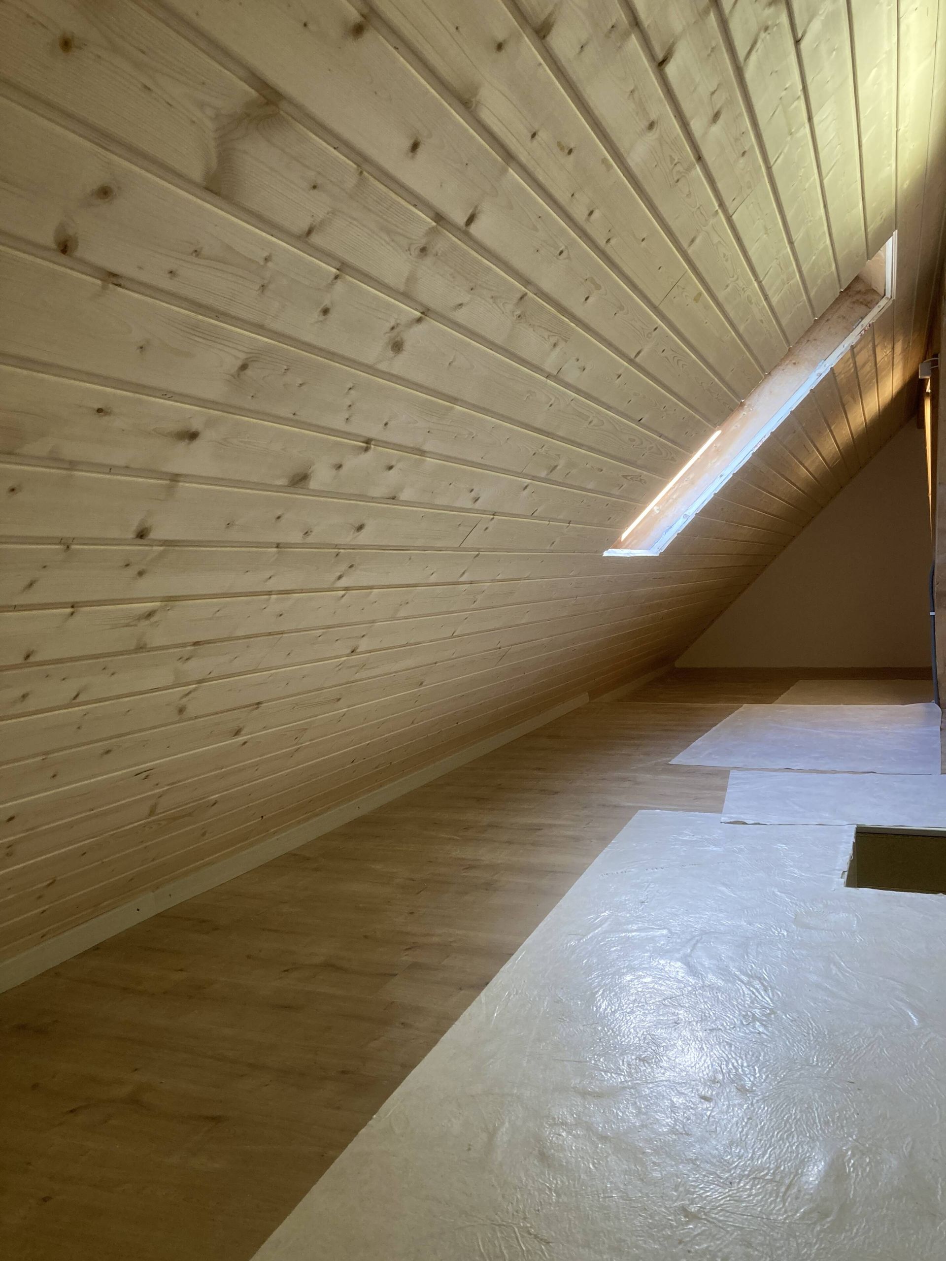 Dachzimmer aus Holz mit Dachschräge und kleinem Fenster, Tageslicht.