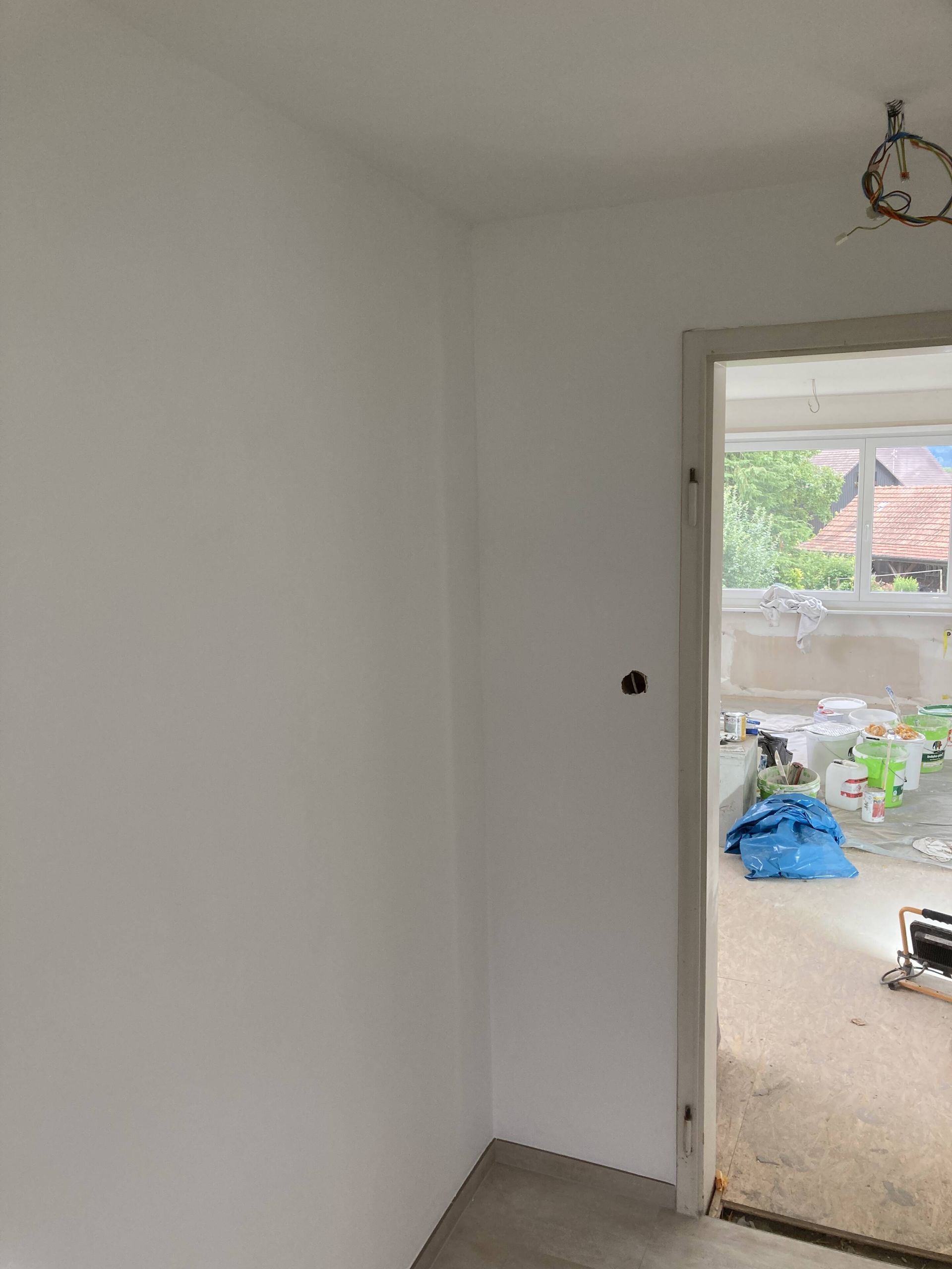 Weiß gestrichene Wände grenzen an einen Türrahmen, dahinter sind Baumaterialien und ein Fenster sichtbar.