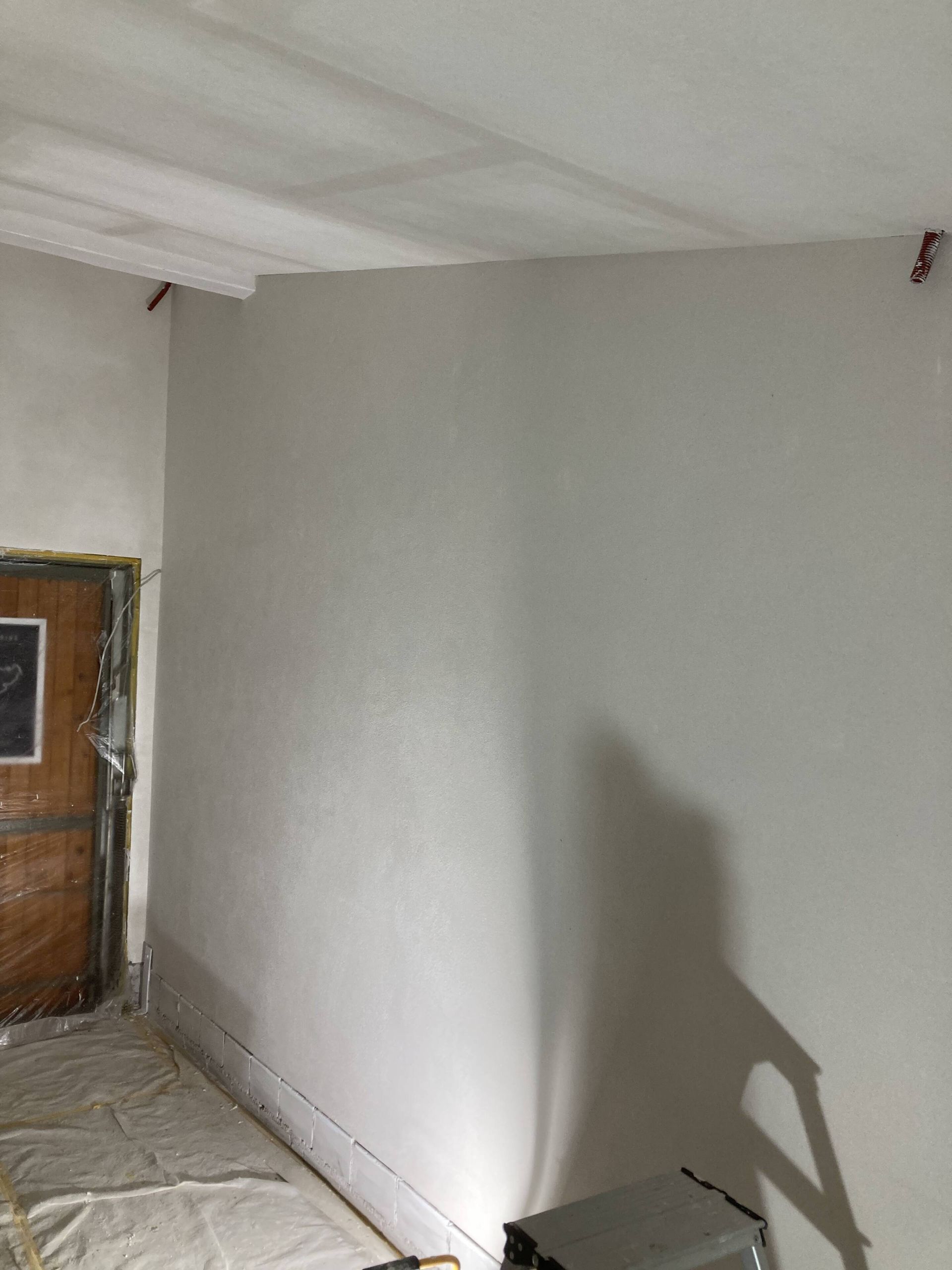 Eine frisch gestrichene, hellgraue Wand und Decke in einem Raum. Eine Holztür und ein Schatten sind sichtbar.