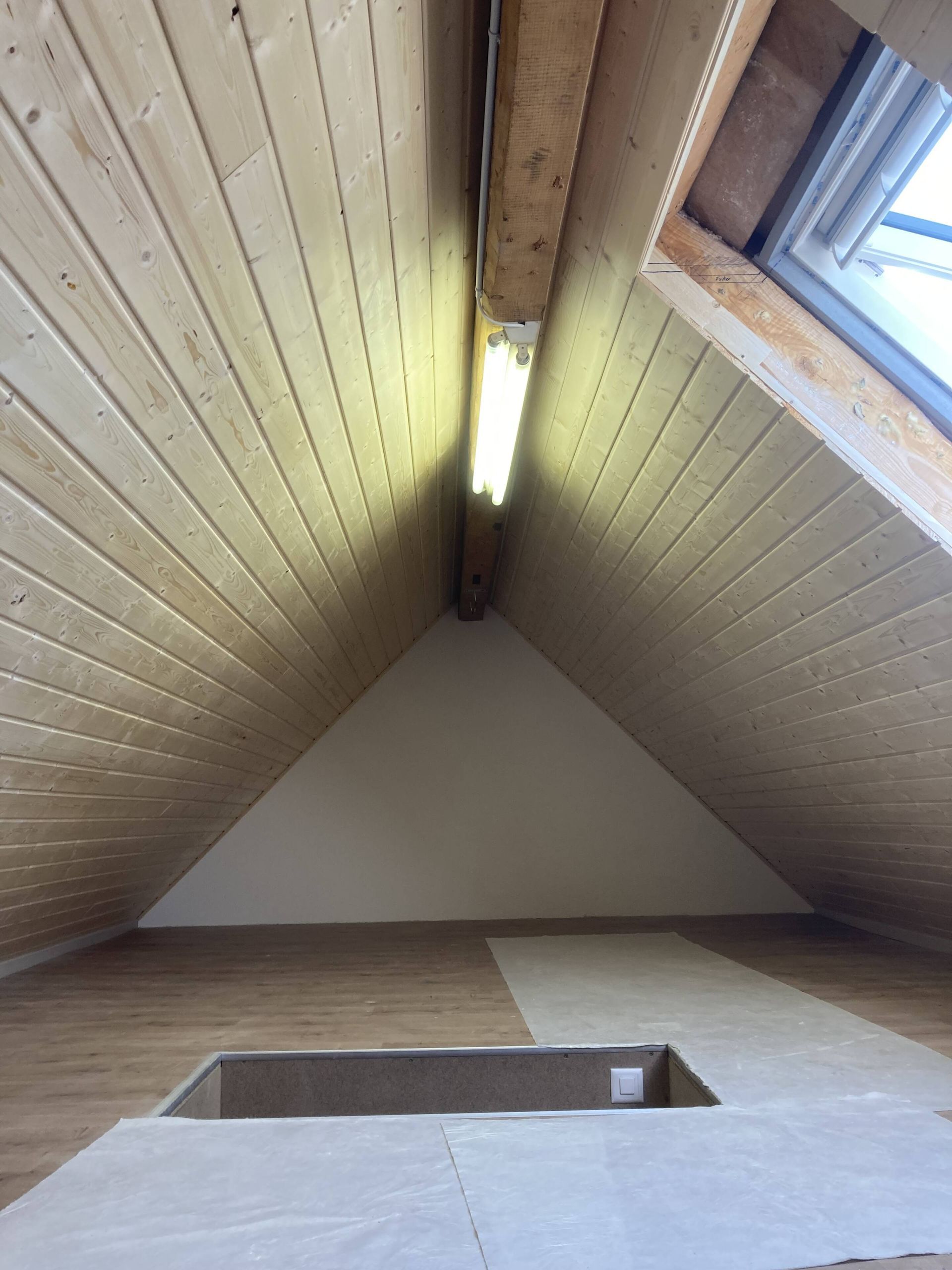 Dachgeschoss mit Holzvertäfelung an der Dachschräge, Oberlicht rechts und rechteckiger Öffnung im Boden.