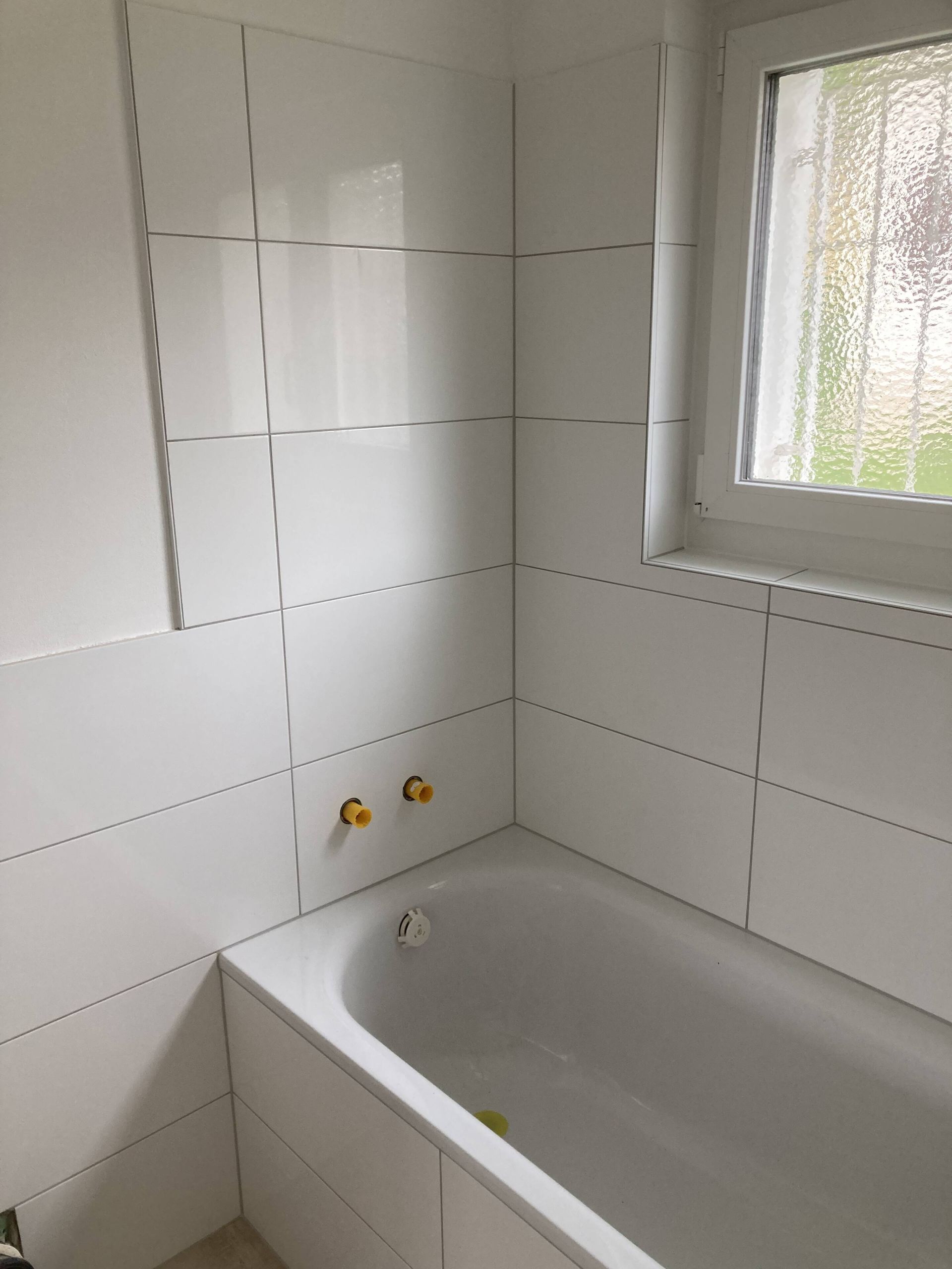 Weiß gefliestes Badezimmer mit Badewanne, goldenem Wasserhahn und kleinem Fenster.