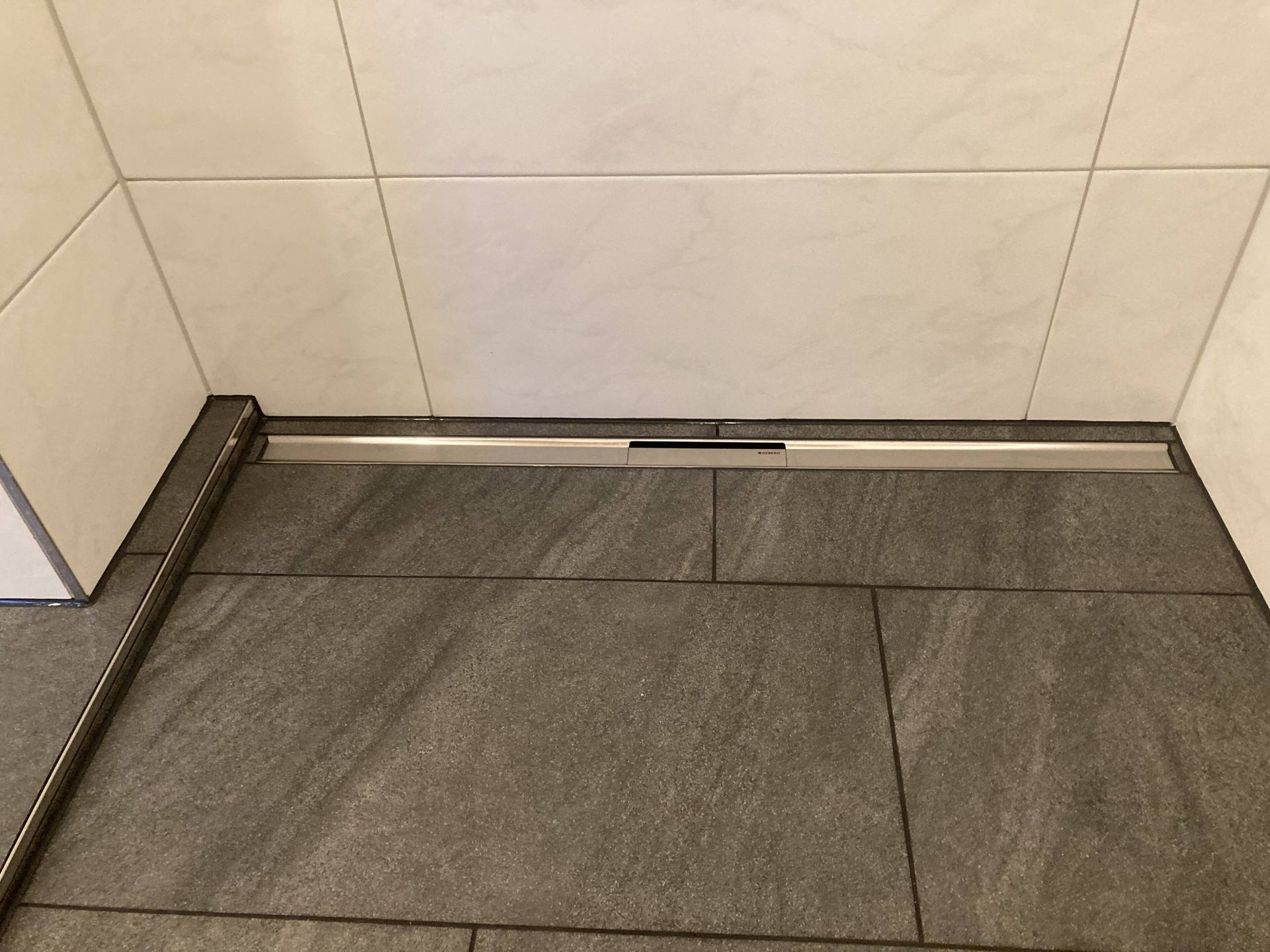 Ein linearer Duschabfluss in einem gefliesten Badezimmer mit grauen Bodenfliesen und weißen Wandfliesen.