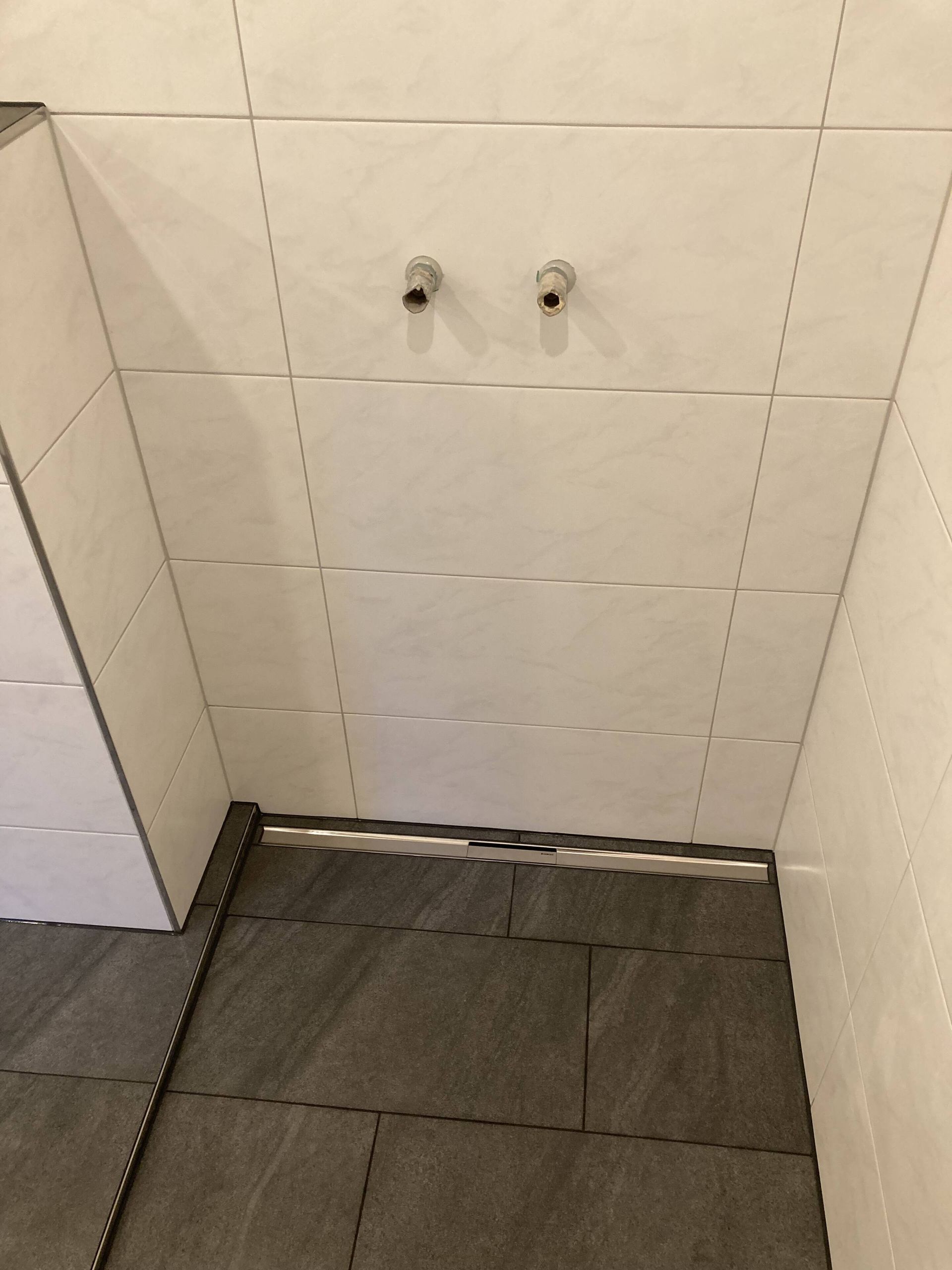 Duschkabine mit weiß gefliesten Wänden, grauen Bodenfliesen, Abfluss und zwei Rohren.