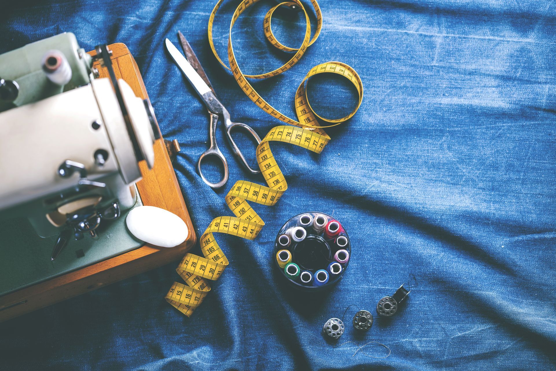 Machine à coudre, ciseaux, mètre ruban et bobines de fil sur du tissu bleu.
