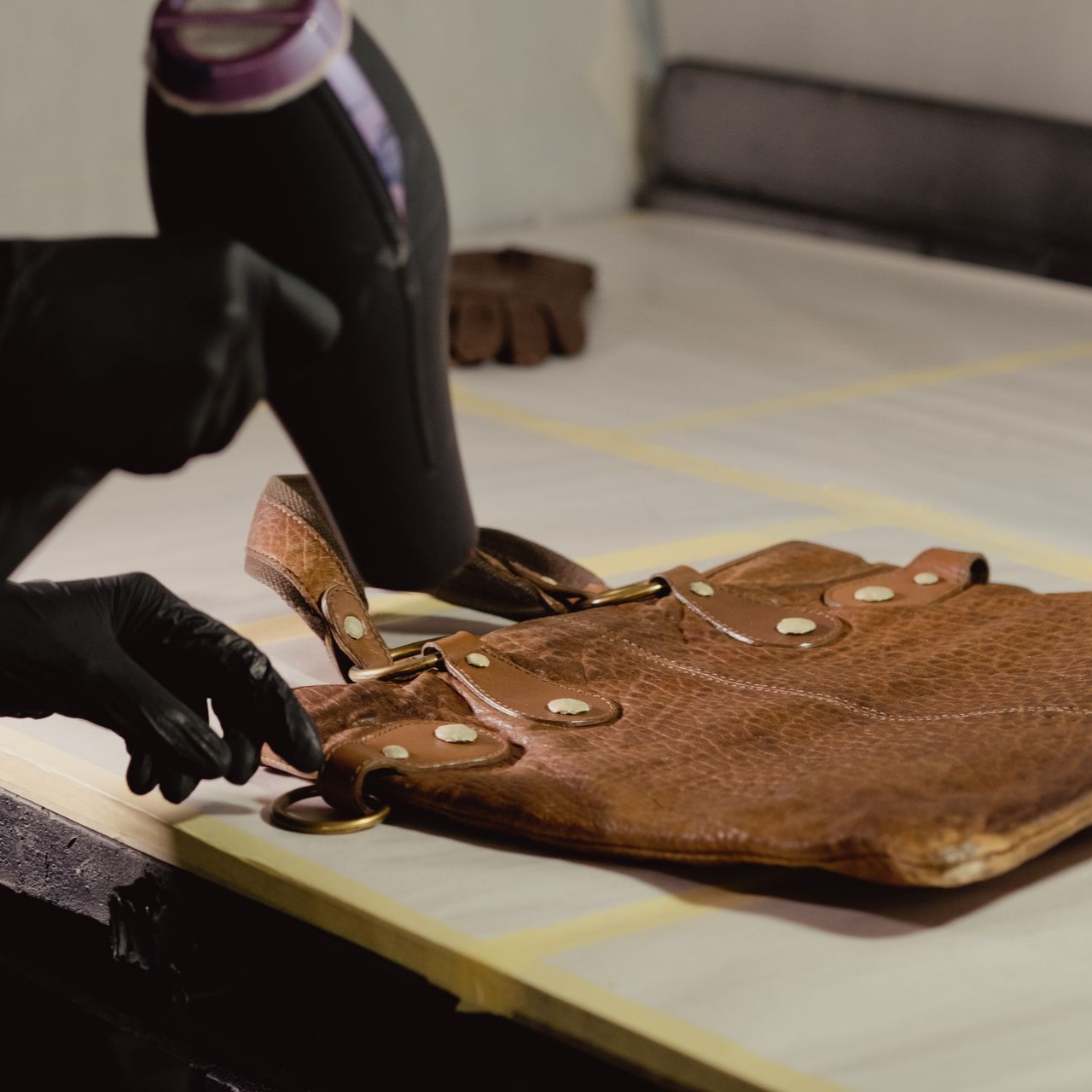 Une personne portant des gants noirs utilise un pistolet thermique sur un sac à main en cuir marron posé sur une table.