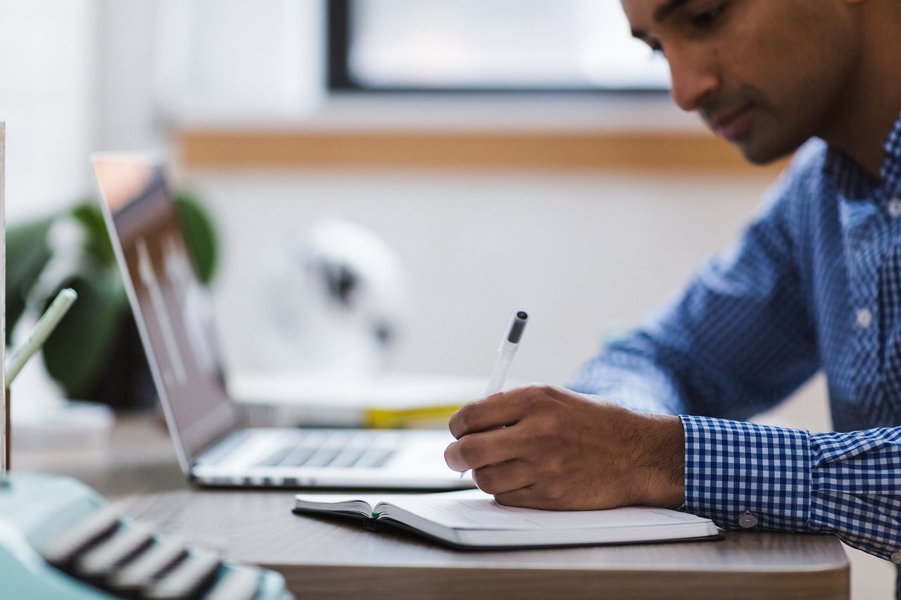 Un hombre está sentado en un escritorio escribiendo en un cuaderno frente a una computadora portátil.