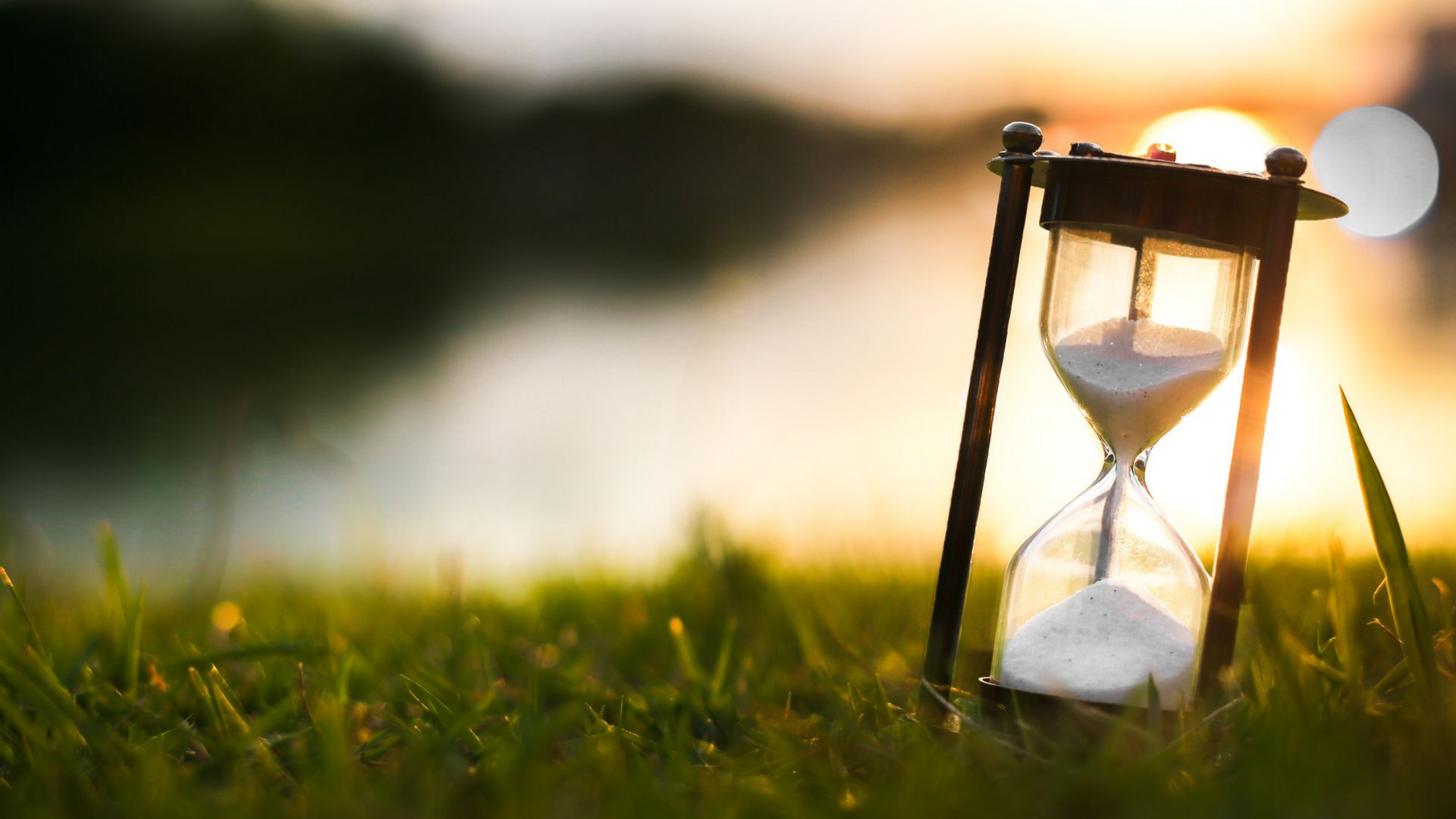 Un sablier sur l'herbe, la lumière du soleil brillant en arrière-plan. Le sable s'écoule, suggérant le passage du temps.