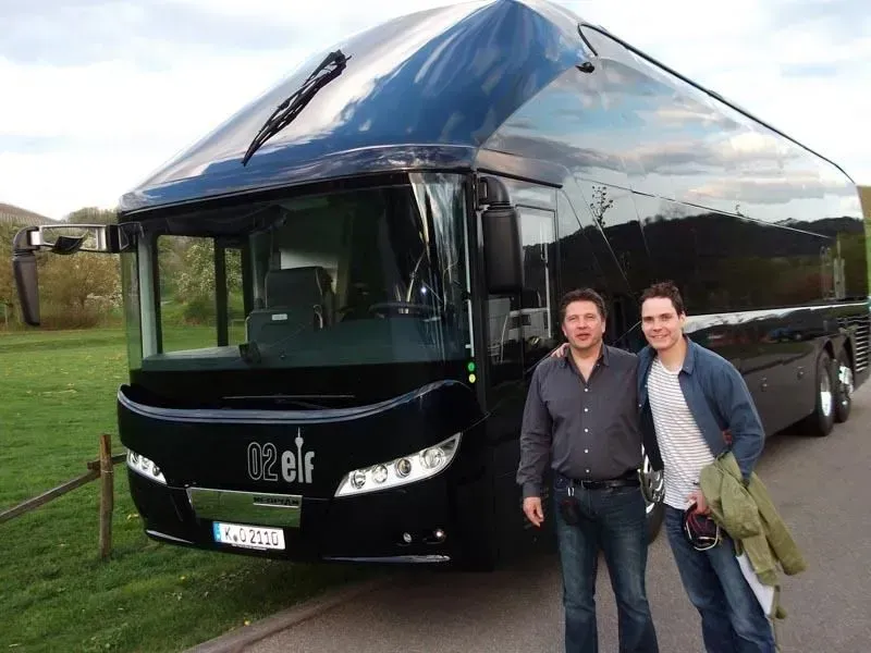 Zwei Männer stehen vor einem schwarzen Bus mit der Aufschrift „Klein“, einer davon Daniel Brühl, 02elf Travel GmbH & Co KG