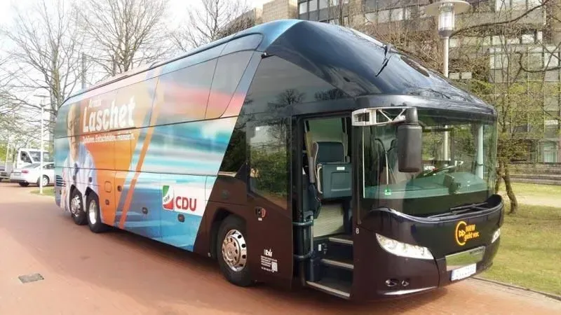 Ein großer blau-oranger Bus steht vor einem Gebäude. 02elf Travel GmbH & Co KG