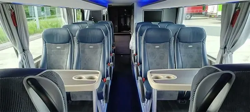Innenansicht eines Reisebusses mit blauen Sitzen und Klapptischen, vom vorderen Gang aus gesehen