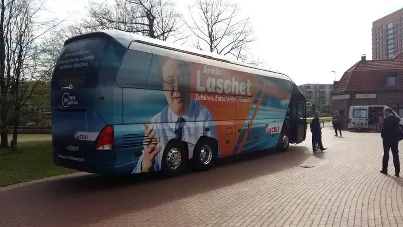 Auf einem gepflasterten Gehweg parkt ein Bus mit dem Bild eines Mannes an der Seite. 02elf Travel GmbH & Co KG