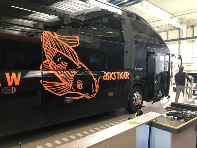 Ein schwarzer Bus mit einem orangefarbenen Fisch an der Seite steht in einer Garage. 02elf Travel GmbH & Co KG