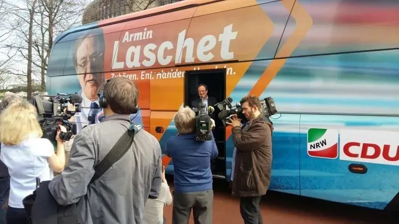 Eine Gruppe von Menschen steht um einen Bus herum, auf dem Armin Laschet steht, 02elf Travel GmbH & Co KG