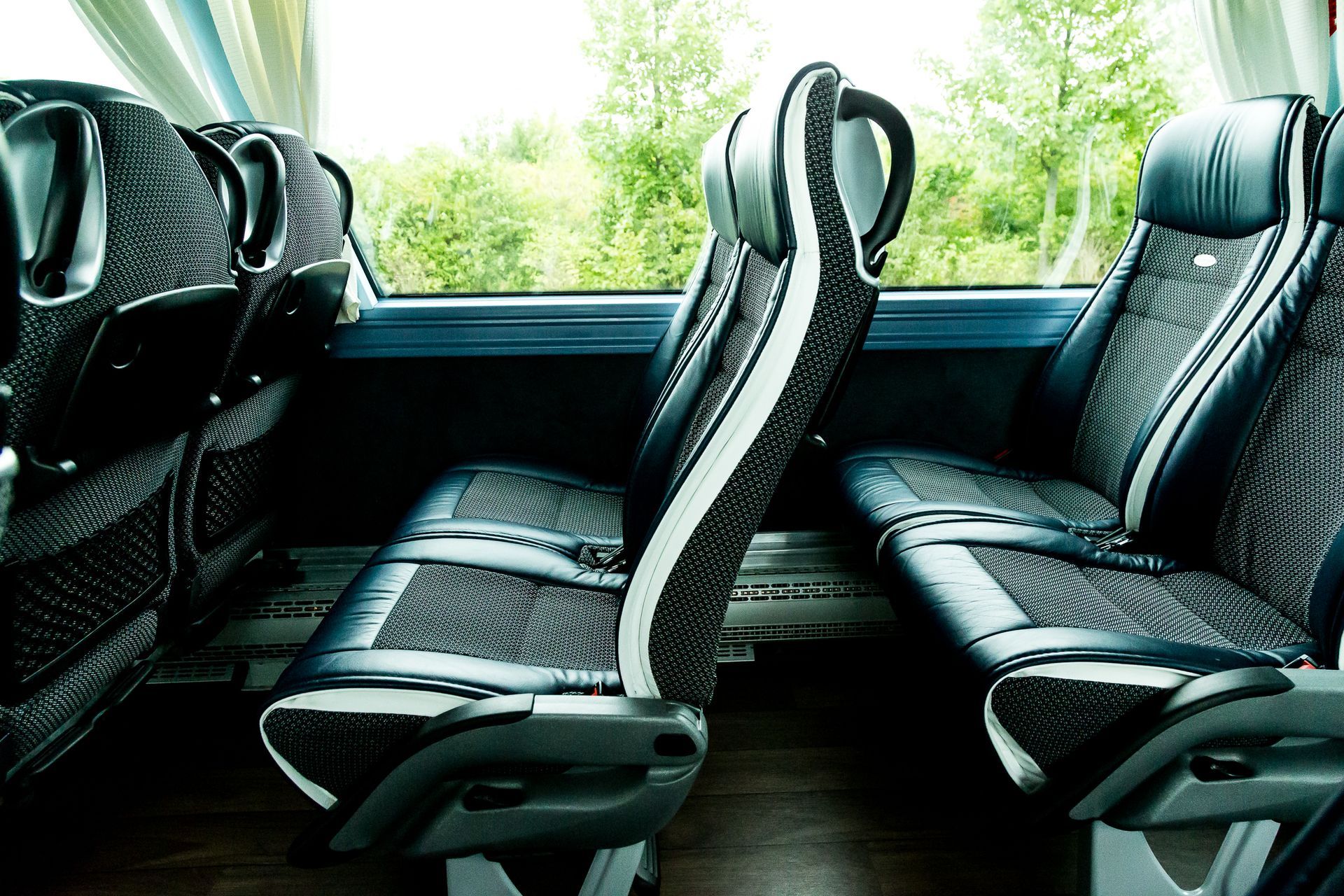 Innenraum eines leeren Busses mit schwarz gemusterten Sitzen und durch die Fenster sichtbaren grünen Bäumen.