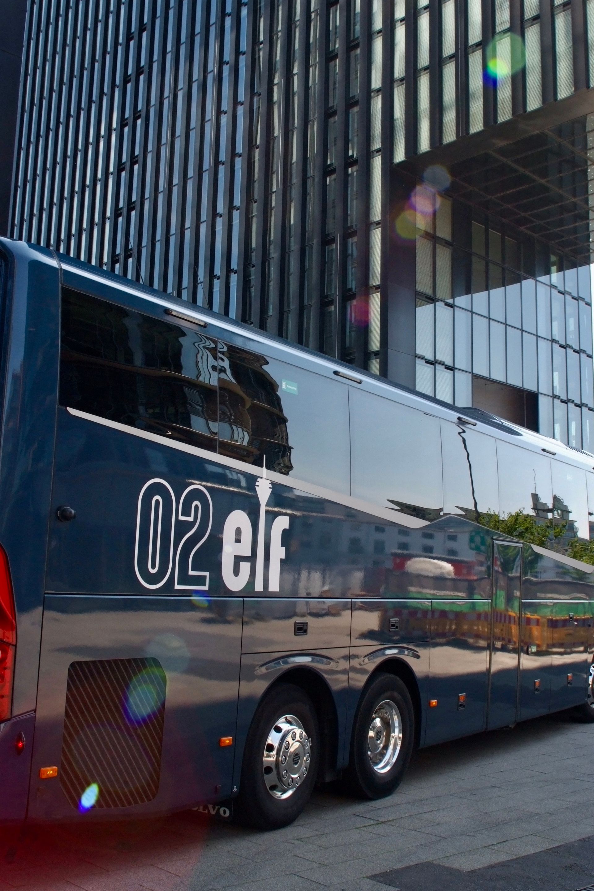 Ein dunkelblauer Doppeldeckerbus mit verspiegelten Fenstern parkt neben einem Glasgebäude.