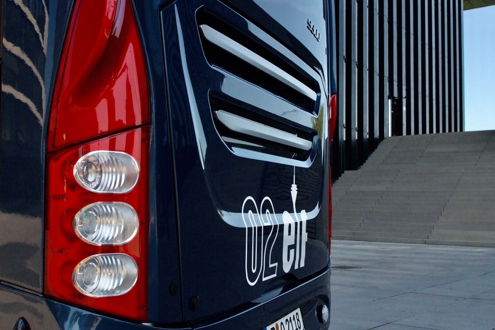 Nahaufnahme eines schwarz-roten Busses mit Rücklichtern und weißer Beschriftung, der in der Nähe eines modernen Gebäudes geparkt ist.