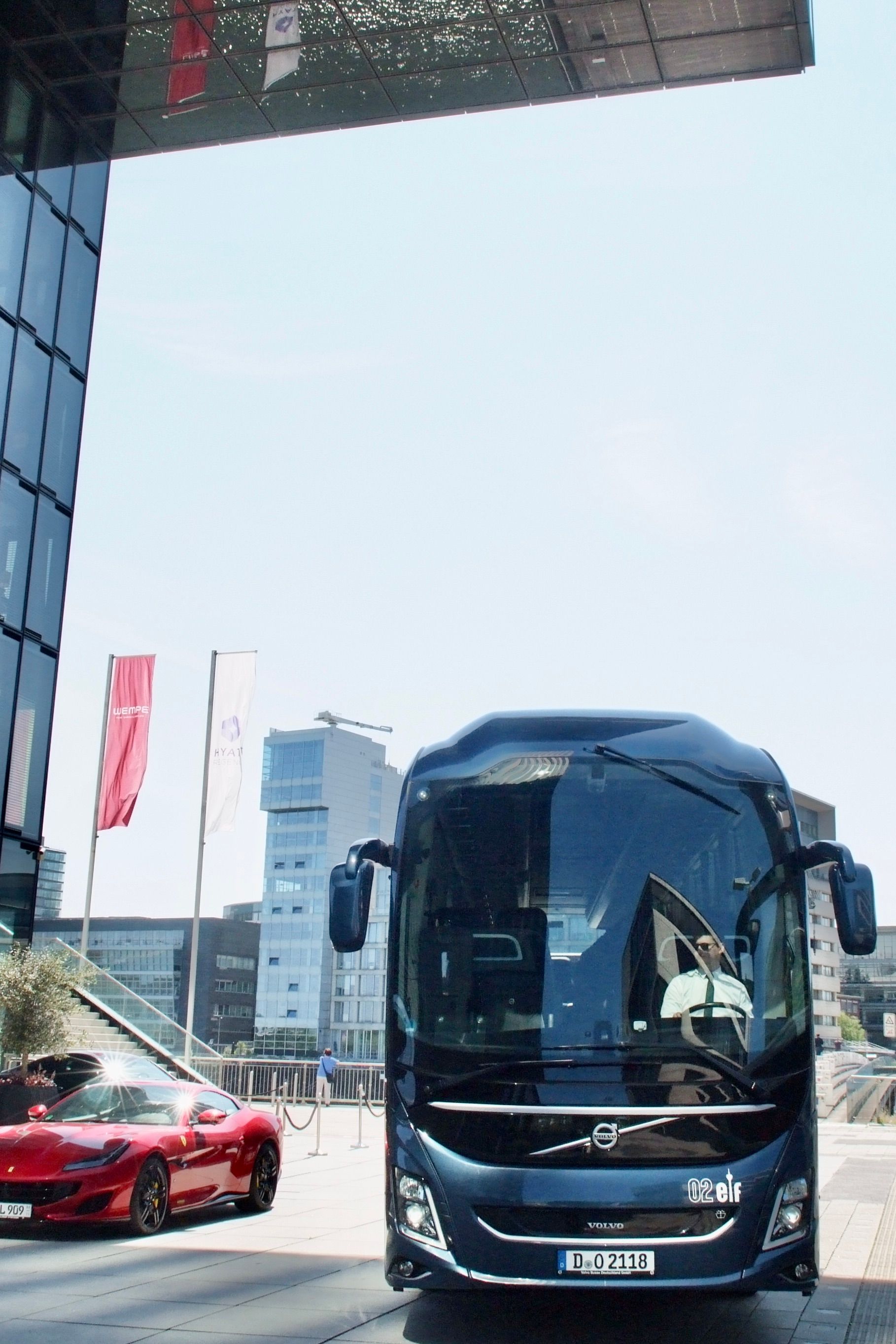 Ein blauer Bus parkt neben einem roten Sportwagen auf einem modernen Stadtplatz unter strahlend blauem Himmel.